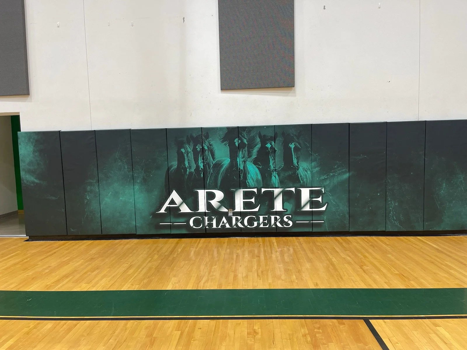 A gymnasium with a wooden floor and a wall padding featuring a graphic of five horses and the words "ARETTE CHARGERS" in large white text.