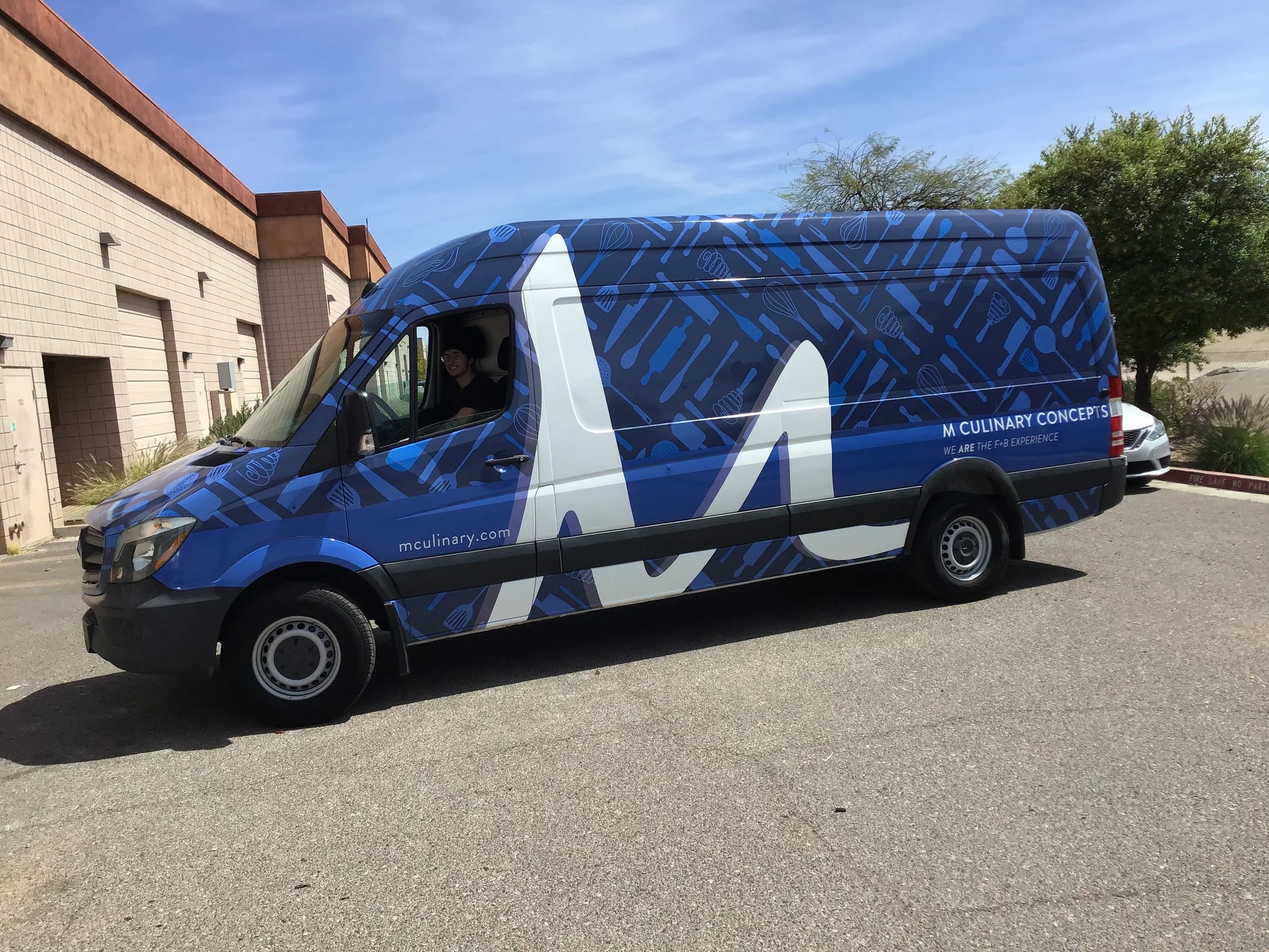 A delivery van with blue and white graphics and text, parked outdoors near a building and trees, with a person inside smiling.
