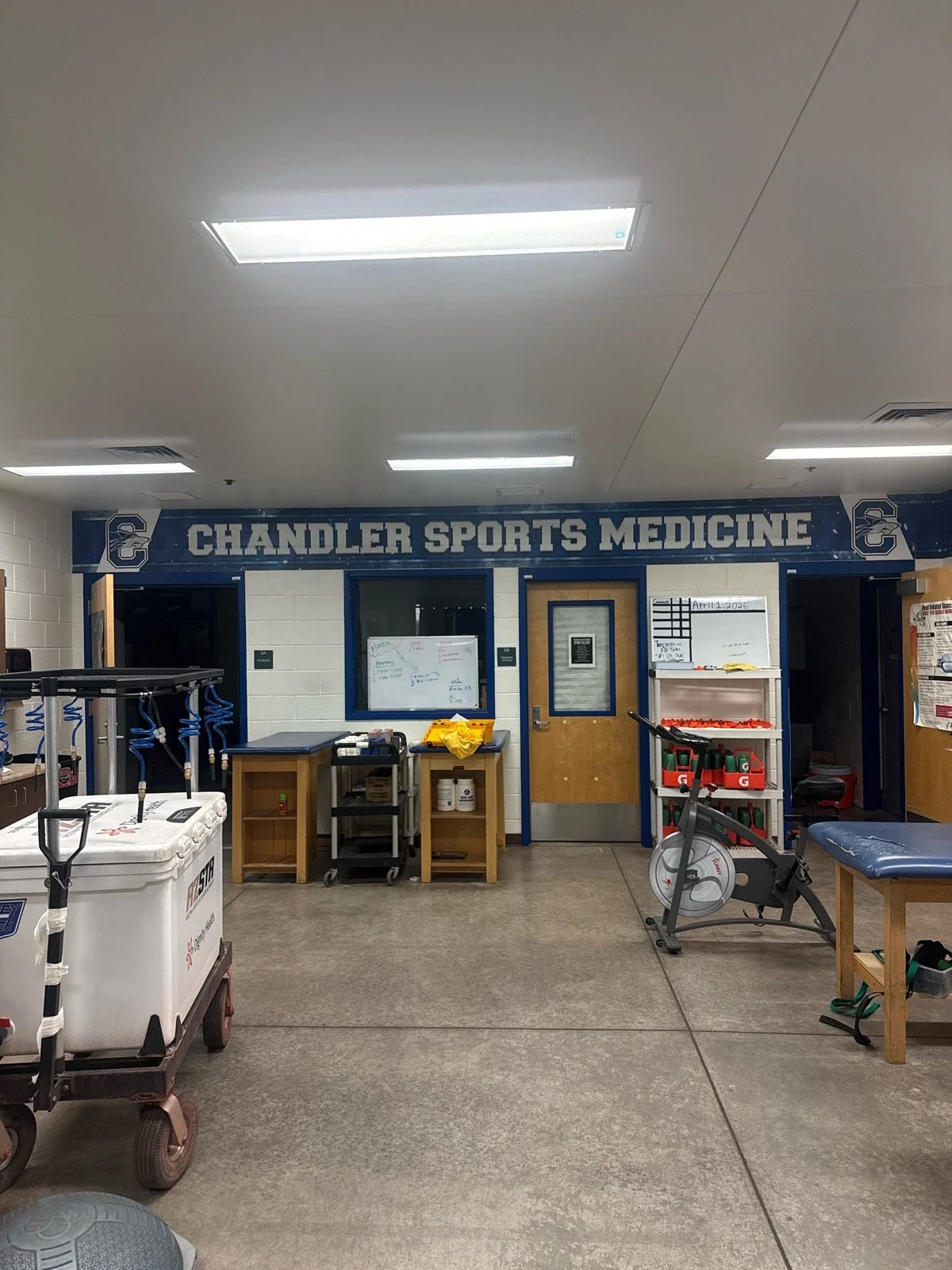 Indoor sports medicine facility with a sign reading 'Chandler Sports Medicine' above the entrance. The room contains equipment for sports therapy, including therapy balls, a stationary exercise bike, and various medical supplies and furniture.