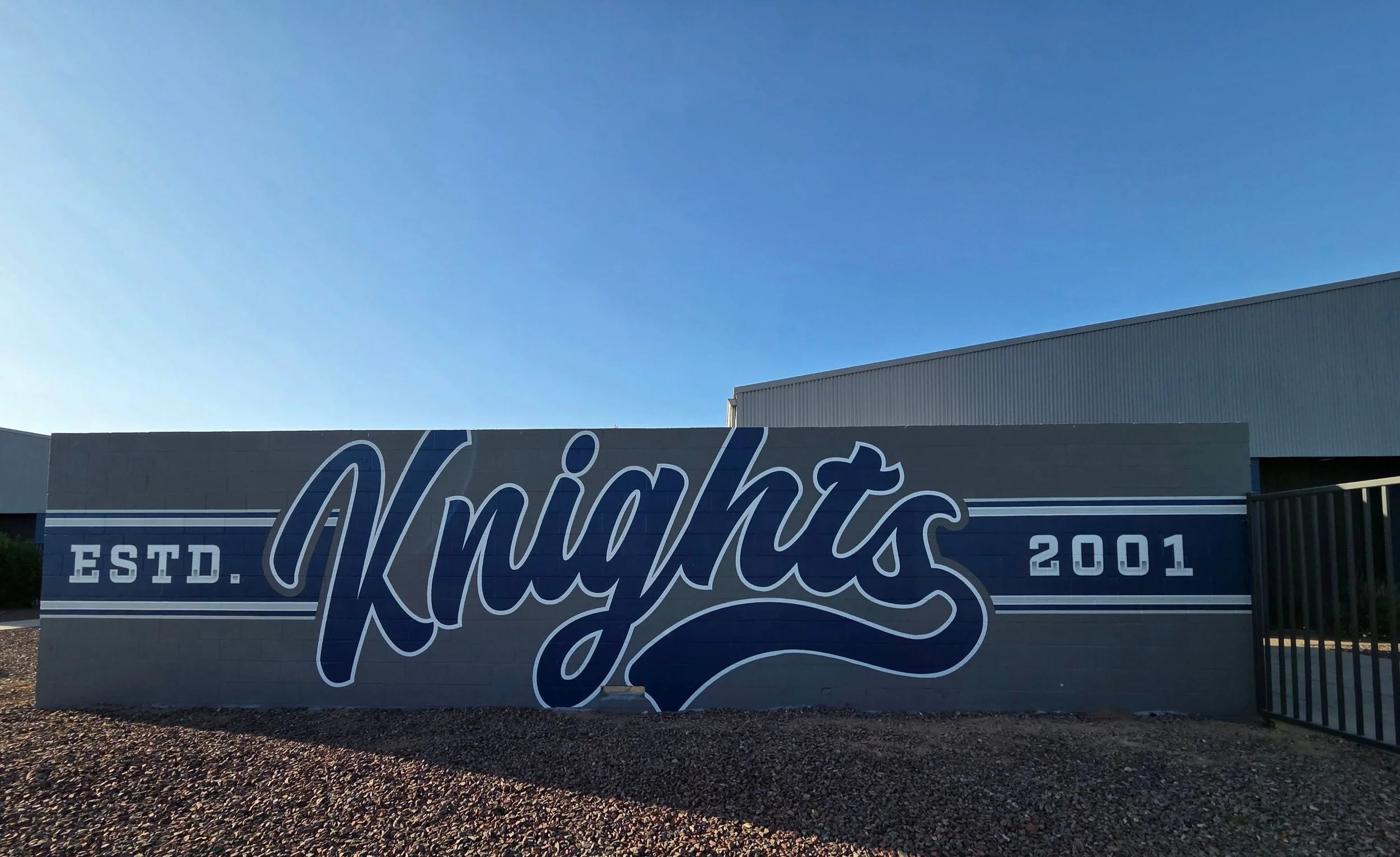 A large blue and white sign on a gray wall reads 'Night's' with 'ESTD. 2001' underneath. The sign is outdoors against a clear blue sky with a gray industrial building partially visible in the background.