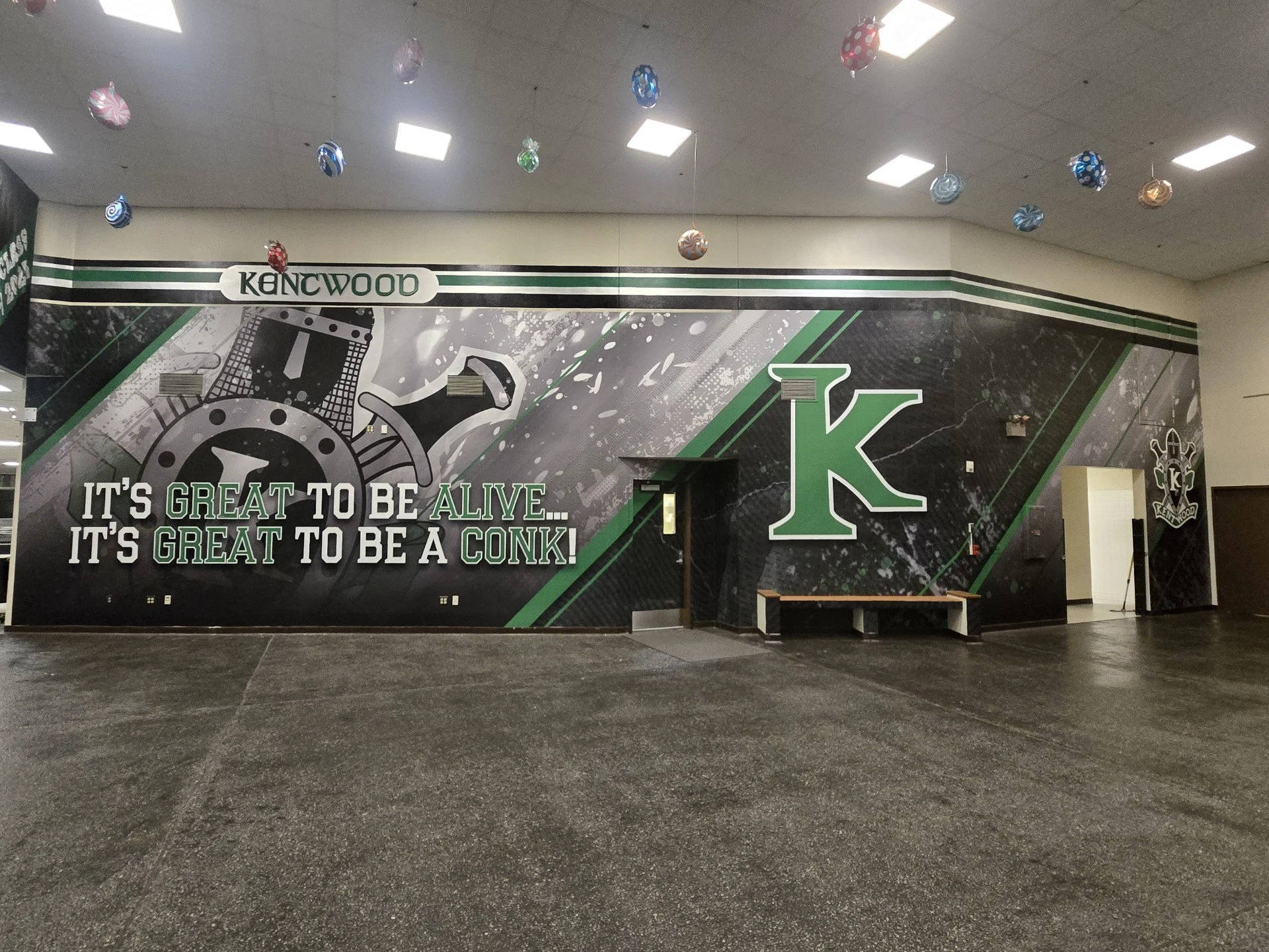 Indoor sports arena with stylized mural featuring a large letter 'K' and a stopwatch design, congratulatory text about being a KONK, and hanging decorative balls from the ceiling.