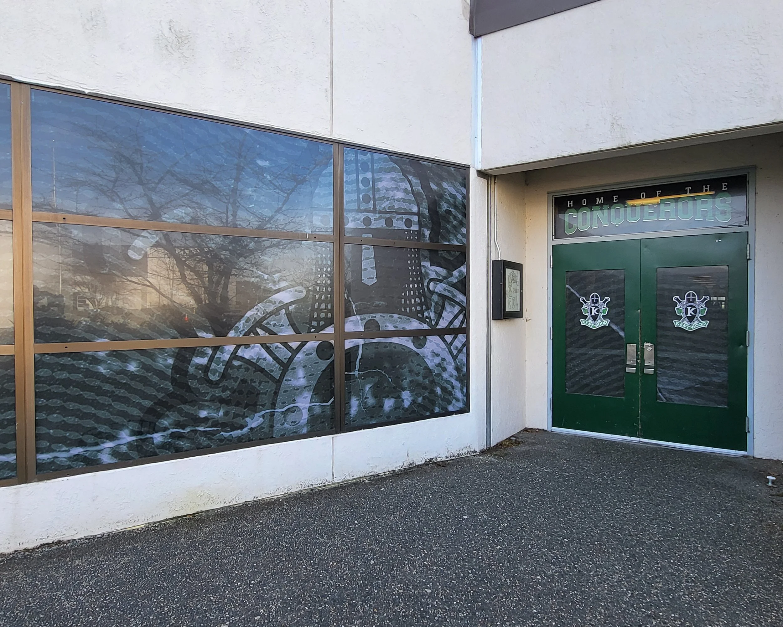 The image shows a storefront with a large glass window reflecting trees and the sky, and a green door with signs reading "Home of the Conquerors" and a logo with a knight's helmet.