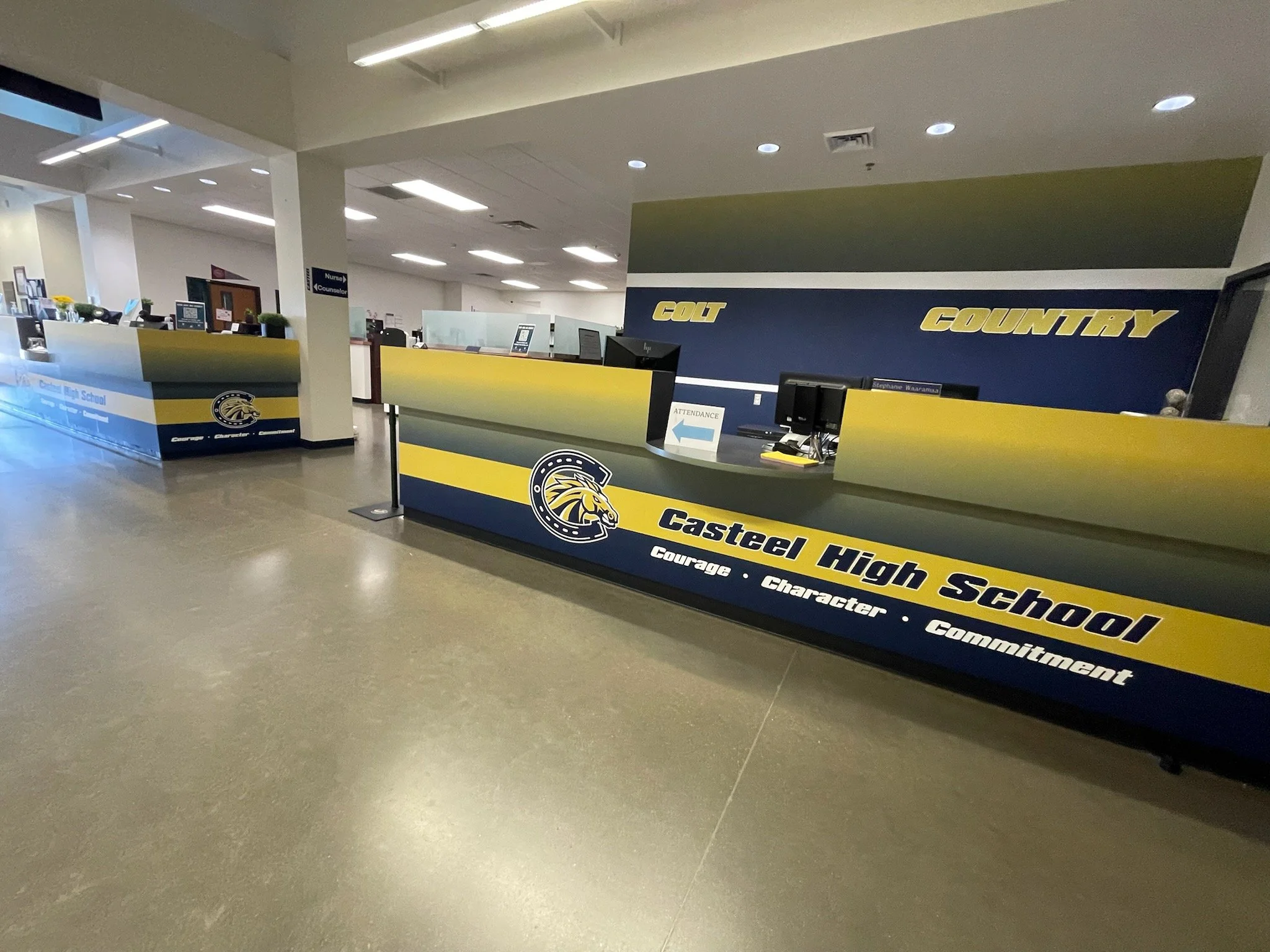 Inside view of a school reception area with a blue and yellow color scheme, featuring a counter with computers and signage for a high school.