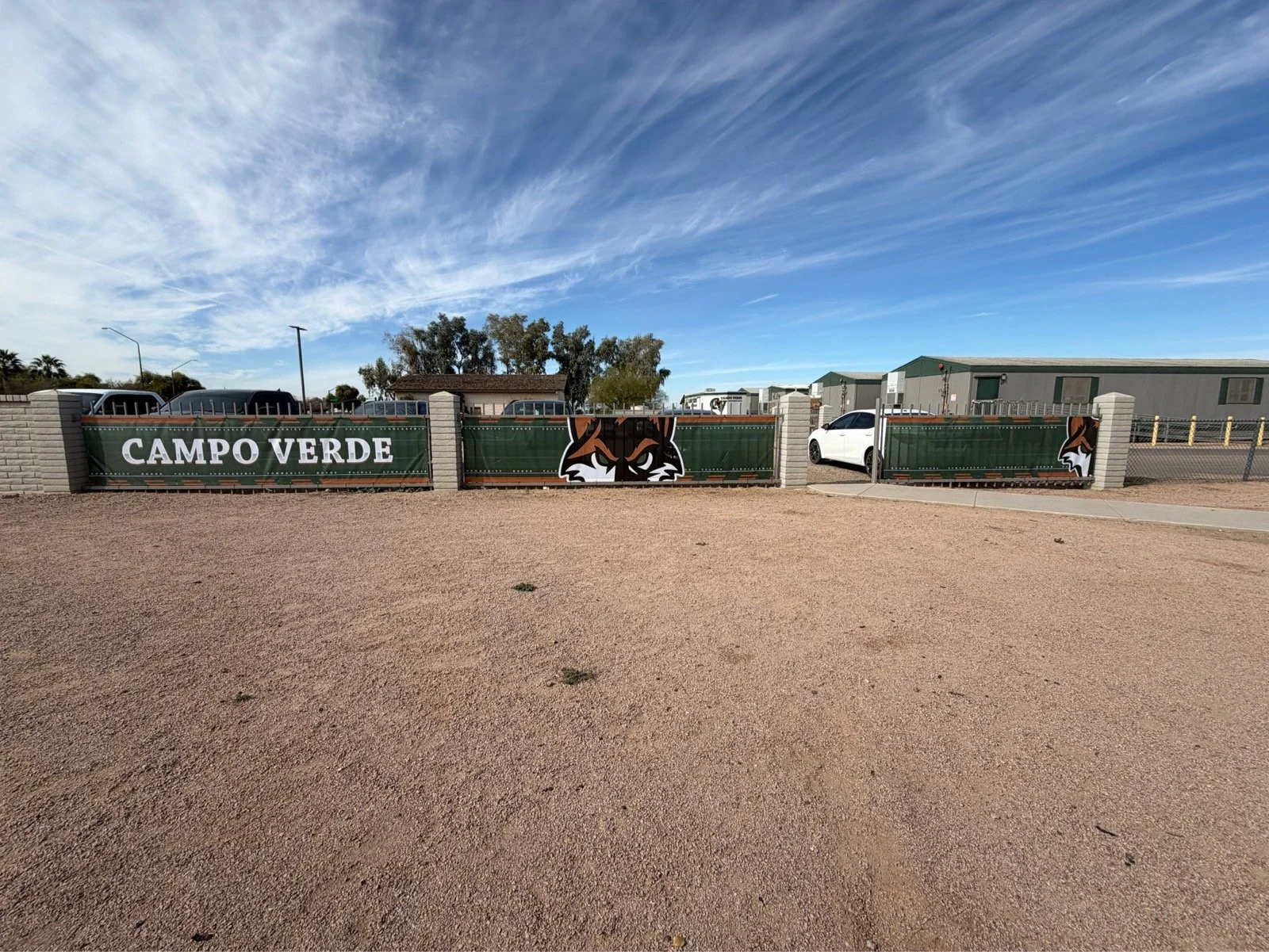A green gate with the words 'CAMPO VERDE' and a stylized fox head logo, set against a background of a parking lot and blue sky with wispy clouds.
