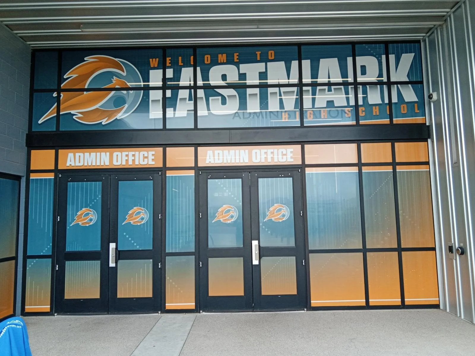 Entrance to Eastmark High School with glass doors and windows displaying school logo, name, and 'Admin Office' signs above the doors.