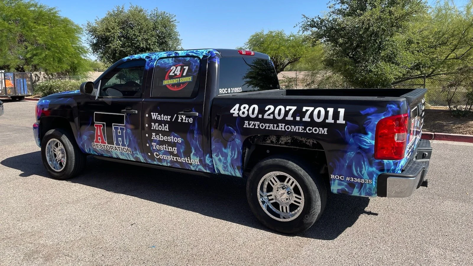 Black pickup truck with blue flame graphics, advertising AH Restoration, listing services like water and fire damage repair, mold removal, asbestos testing, and construction, with contact info and website, parked on a paved surface with trees in the 