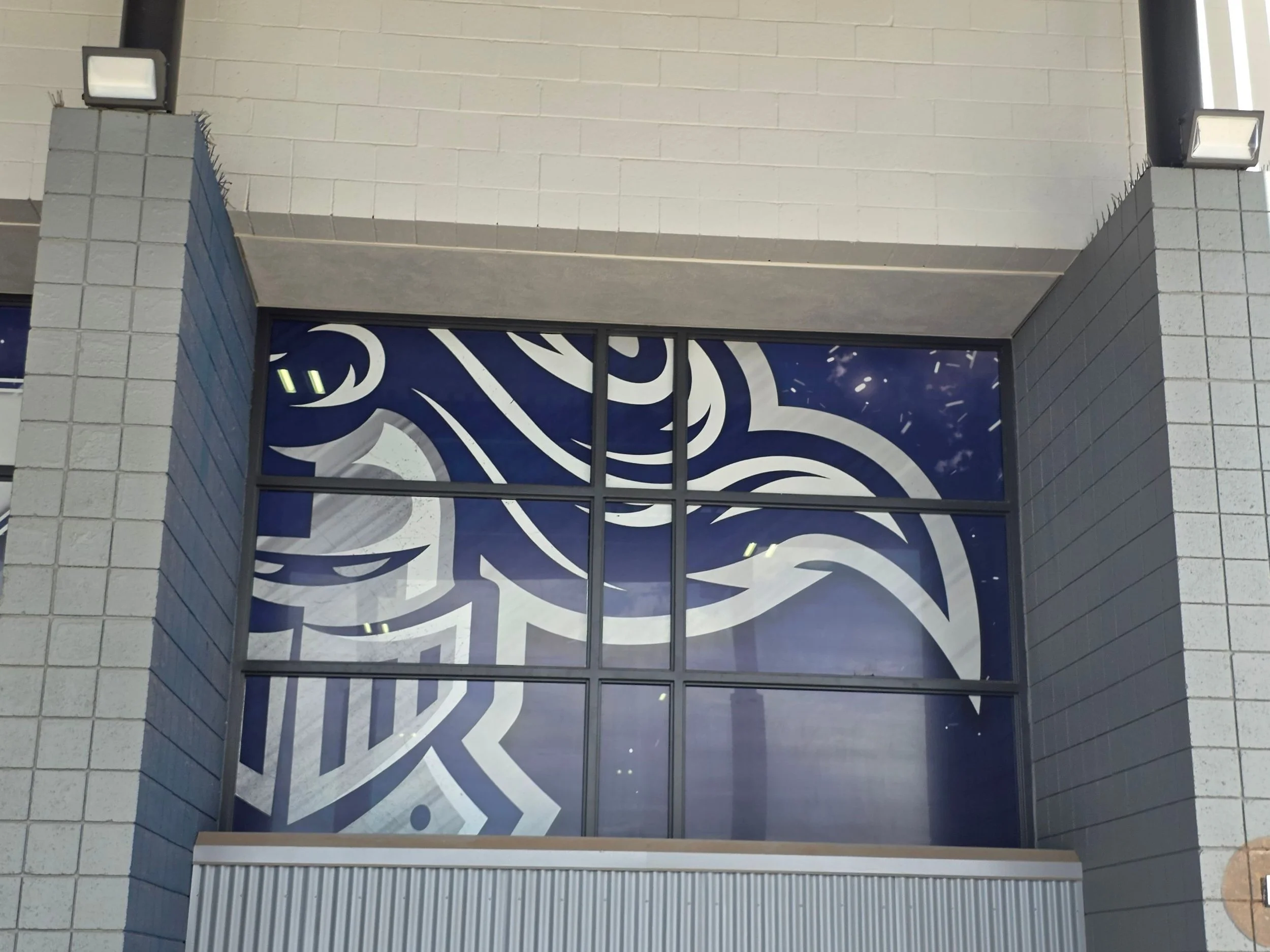 The exterior of a building featuring large glass windows with a swirling pattern and gray brick columns on either side, flanked by two black and white wall-mounted lights.