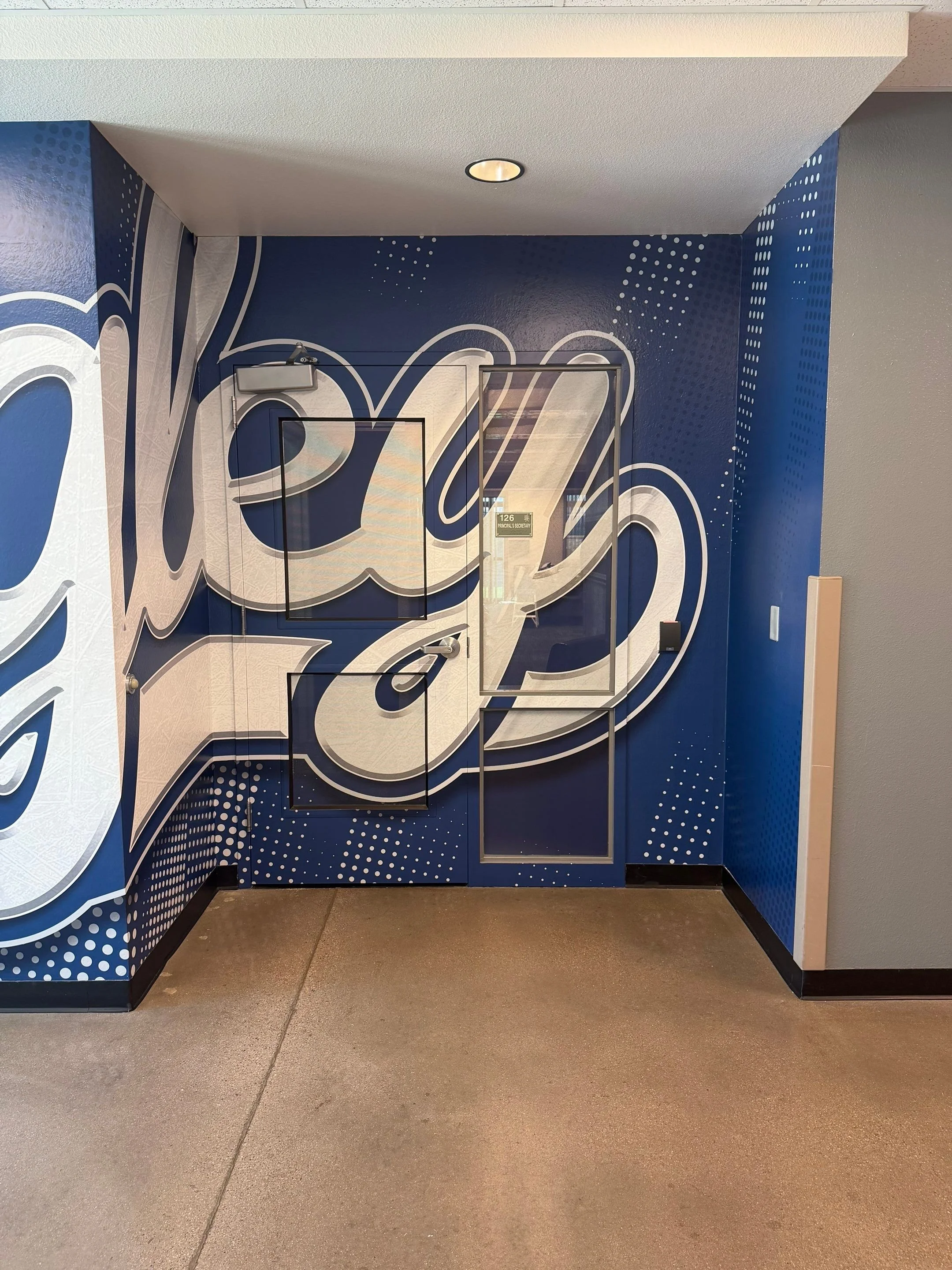 Blue wall with white and gray graffiti-style artwork, including letter designs, and a door with a small window in an indoor hallway.