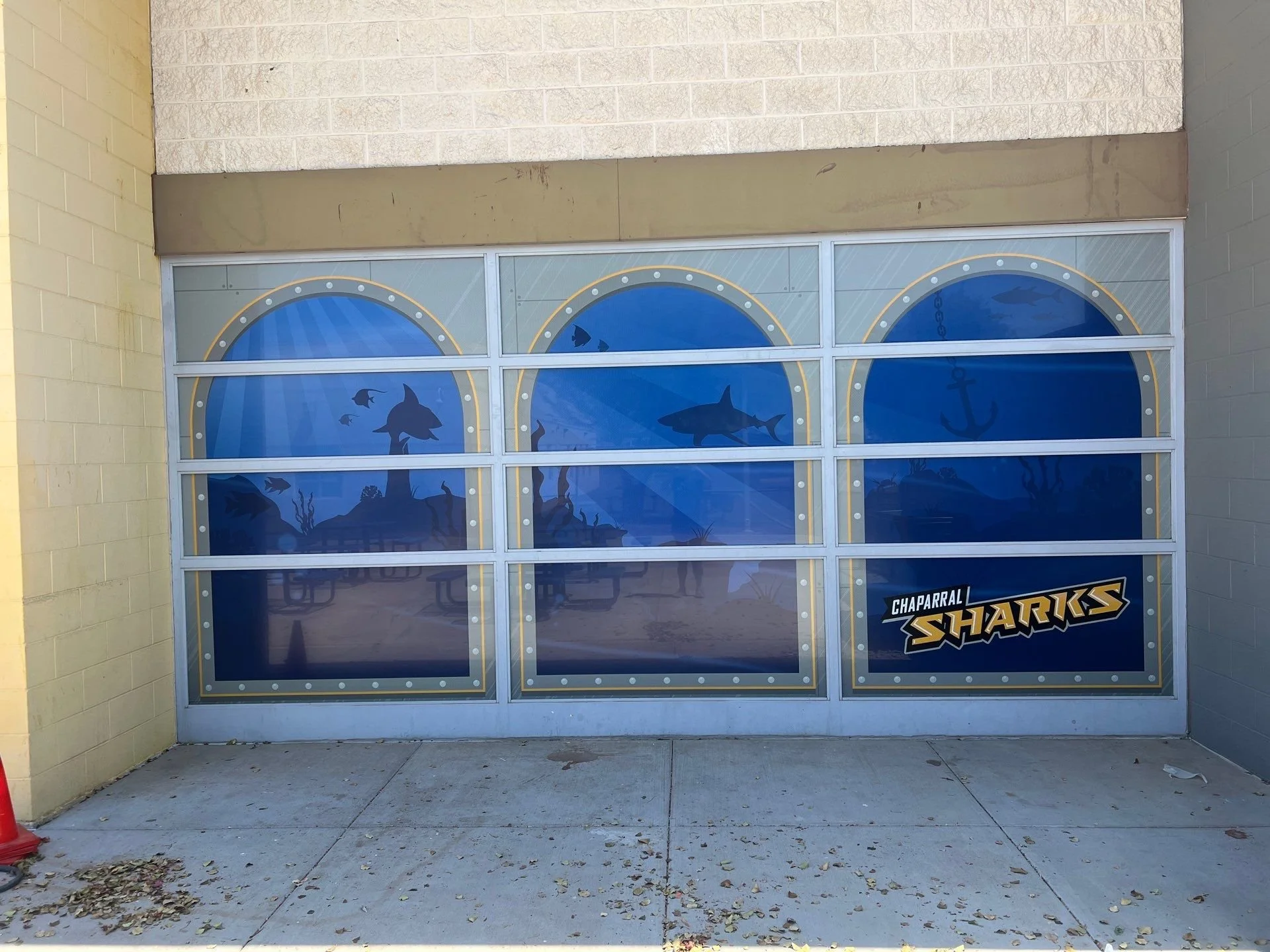 Storefront window with a blue aquatic themed decal featuring fish, an anchor, and underwater plants, with a sign that reads 'Chaparral Sharks.'