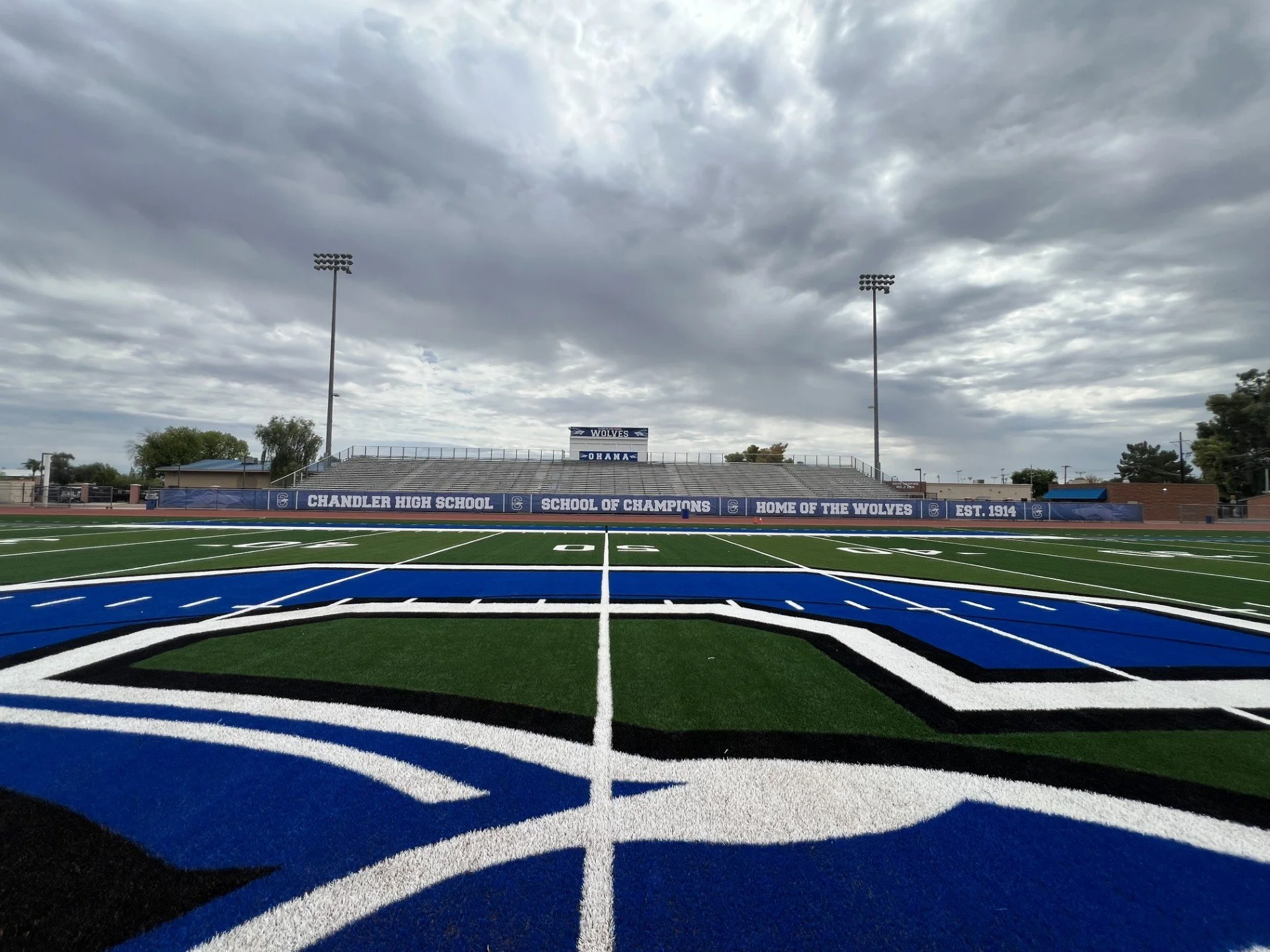 View of a football field with vibrant blue and black end zone markings and white yard lines, surrounded by stadium bleachers with banners reading 'Chandler High School,' 'School of Champions,' 'Home of the Wolves,' and 'Est. 1914.' Overcast sky with 