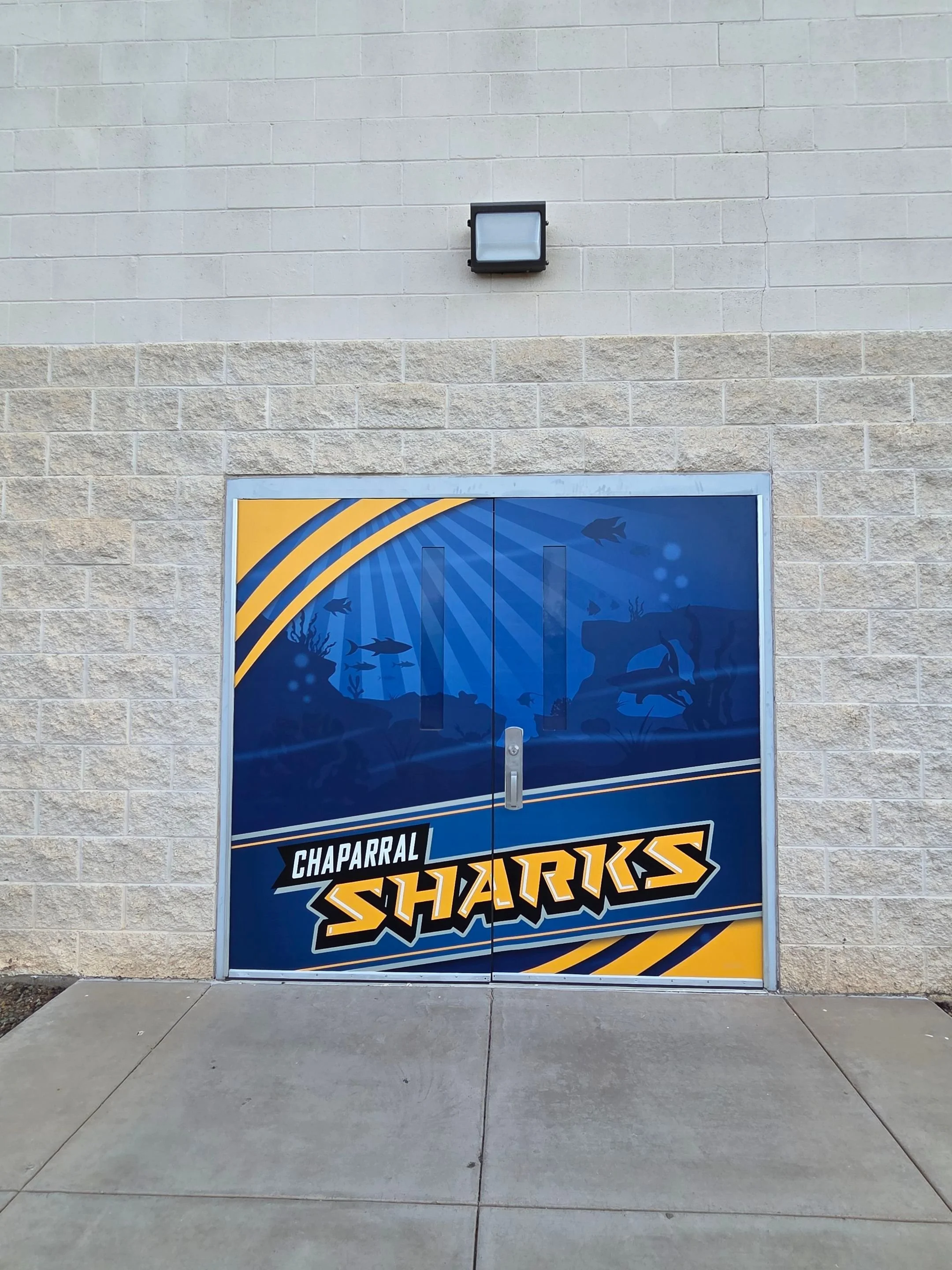 A glass door with a sign for the stuffed animal display, featuring the text 'Chaparral Sharks' and a shark mascot, set in front of a brick wall with a light above it.