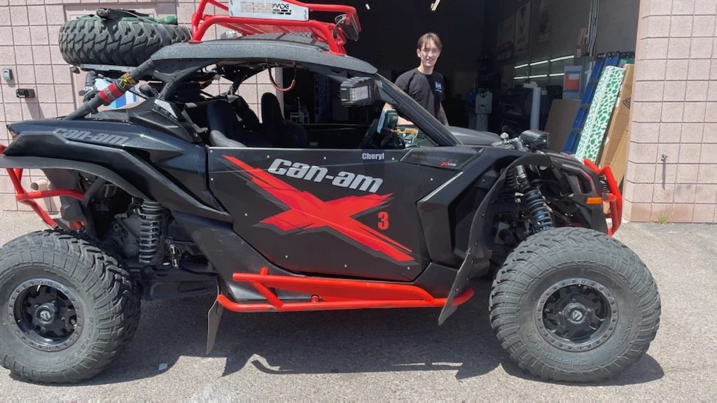 Off-road Can-Am X3 vehicle parked outside a building with a person standing near the driver side, in front of some stacked items and building wall.