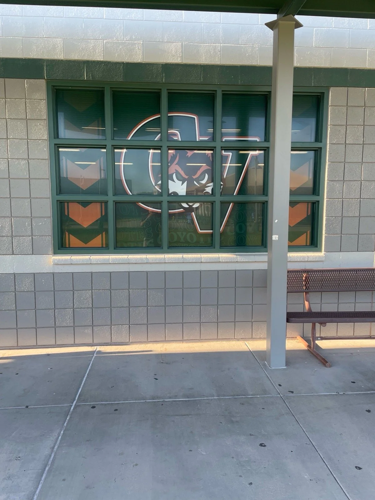 A window with a Michigan State Spartans logo featuring a tiger's face in the center, surrounded by a rectangular frame, on a gray tiled building wall. A bench and a pole are in front of the window.