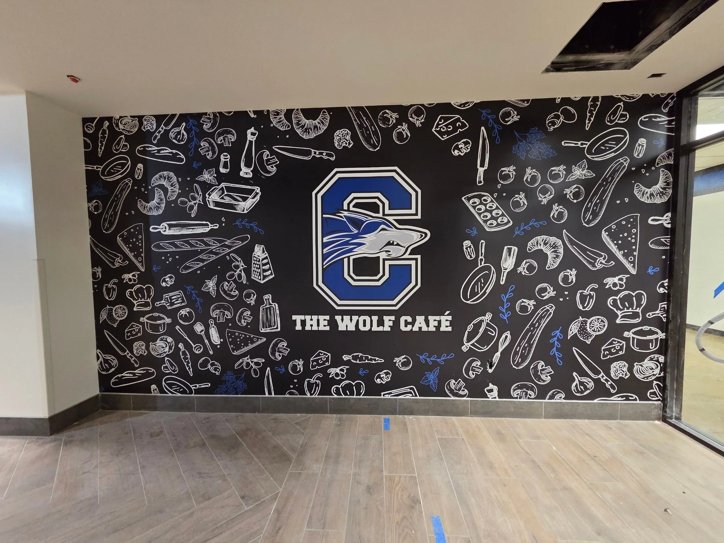 A large black wall with a blue and white logo of a wolf and the text "The Wolf Café" below it, decorated with white line drawings of food items and kitchen utensils.
