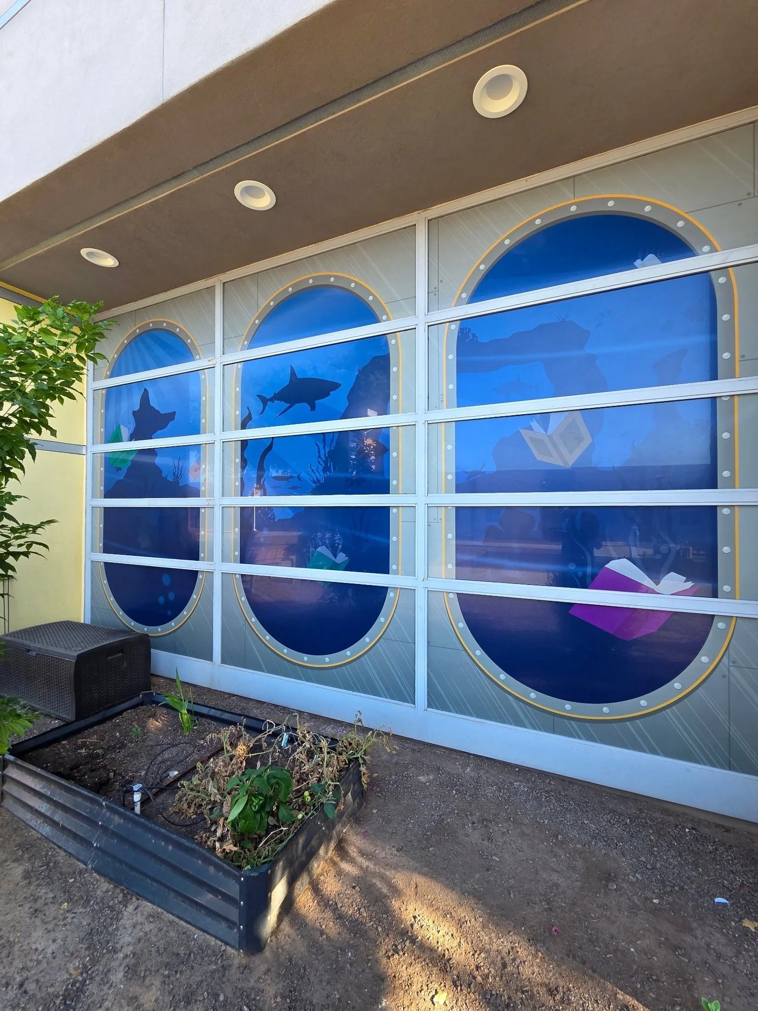 Outdoor view of a building with large windows featuring ocean-themed artwork of fish and marine life; a planter with greenery and a black drainage grate are in the foreground.