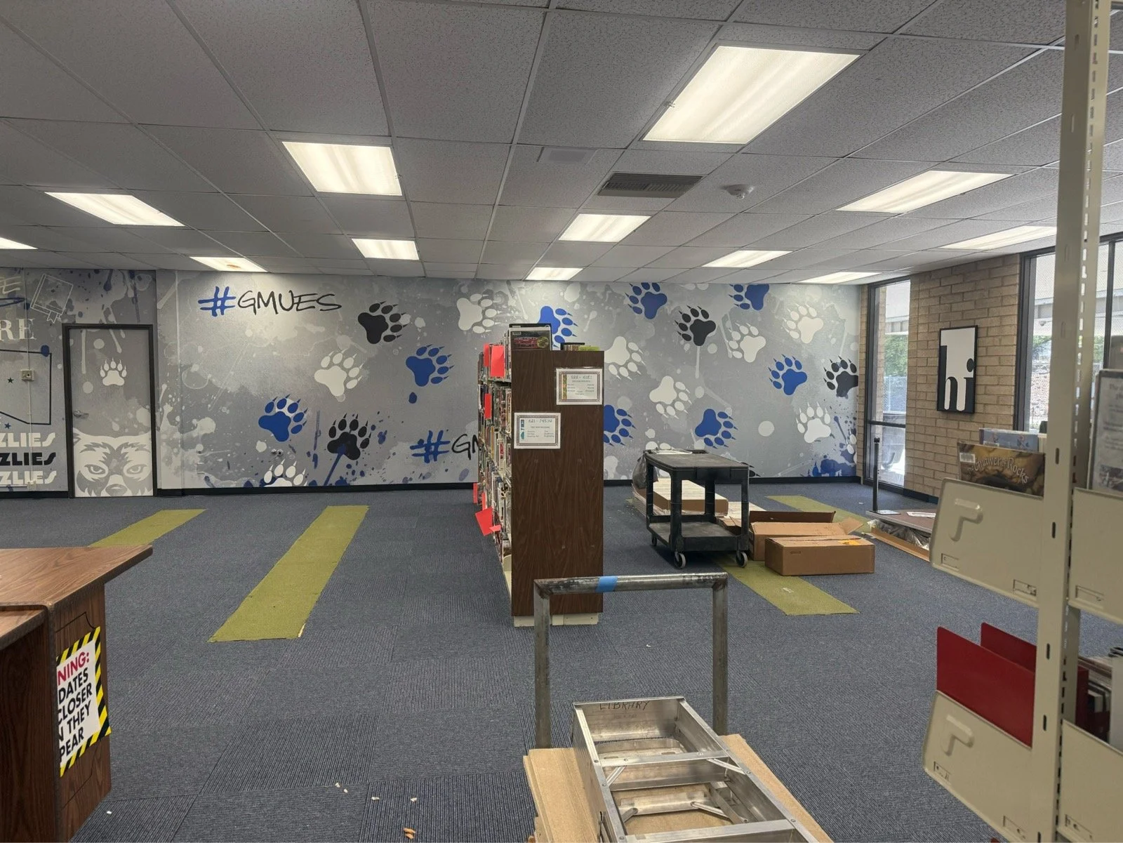 Empty library interior with a gray carpet, a mural wall with paw prints and hashtags, several cardboard boxes, a small table, and a library cart. Part of a sign on the left warns about dates and deadlines.