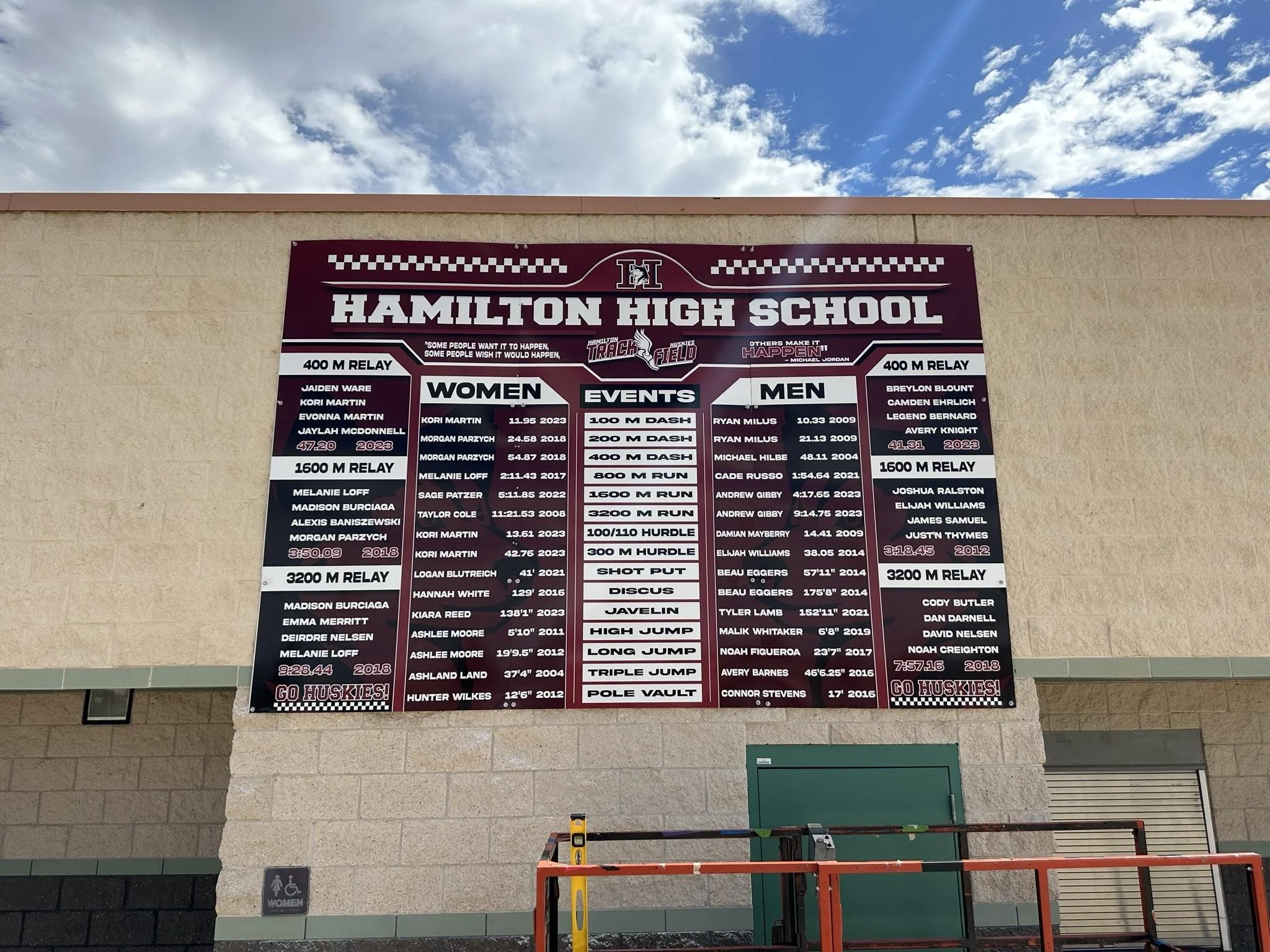 Hamilton High School track records and achievements poster on school wall with a cloudy sky above.