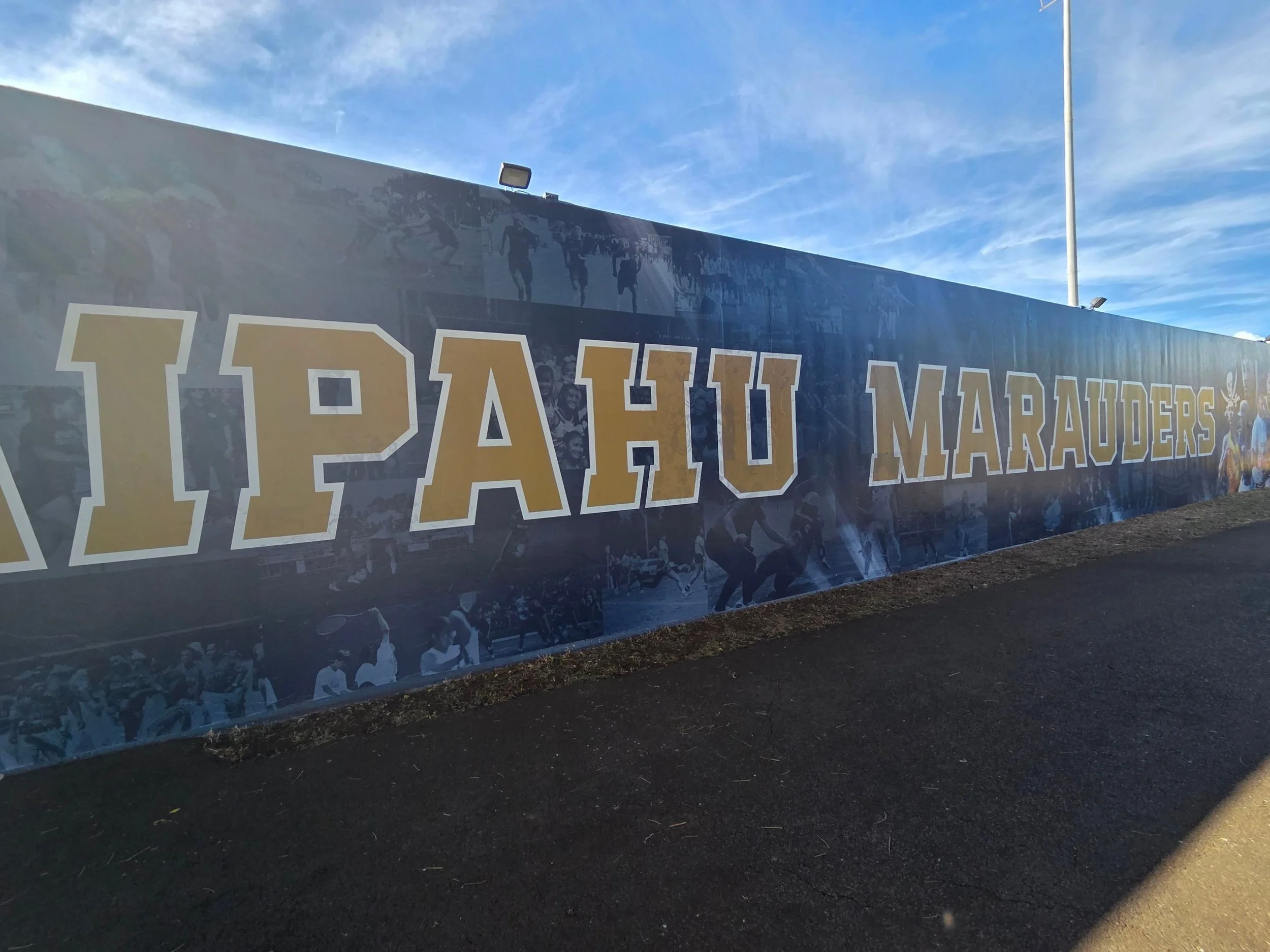 A large outdoor wall with the words 'Paoli Marauders' in yellow and white letters, featuring images of sports players and a football, under a blue sky with some clouds.