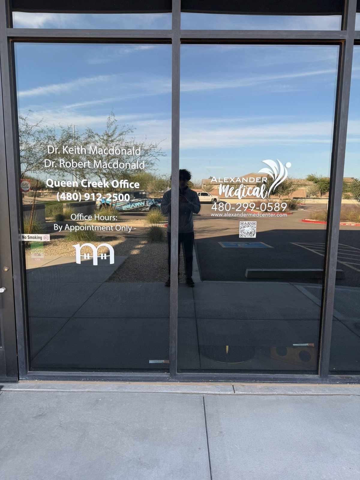 The glass door displays contact and office information for Alexander Medical, including names of doctors, phone number, website, and office hours, with a reflection of the person taking the photo and outdoor scenery.