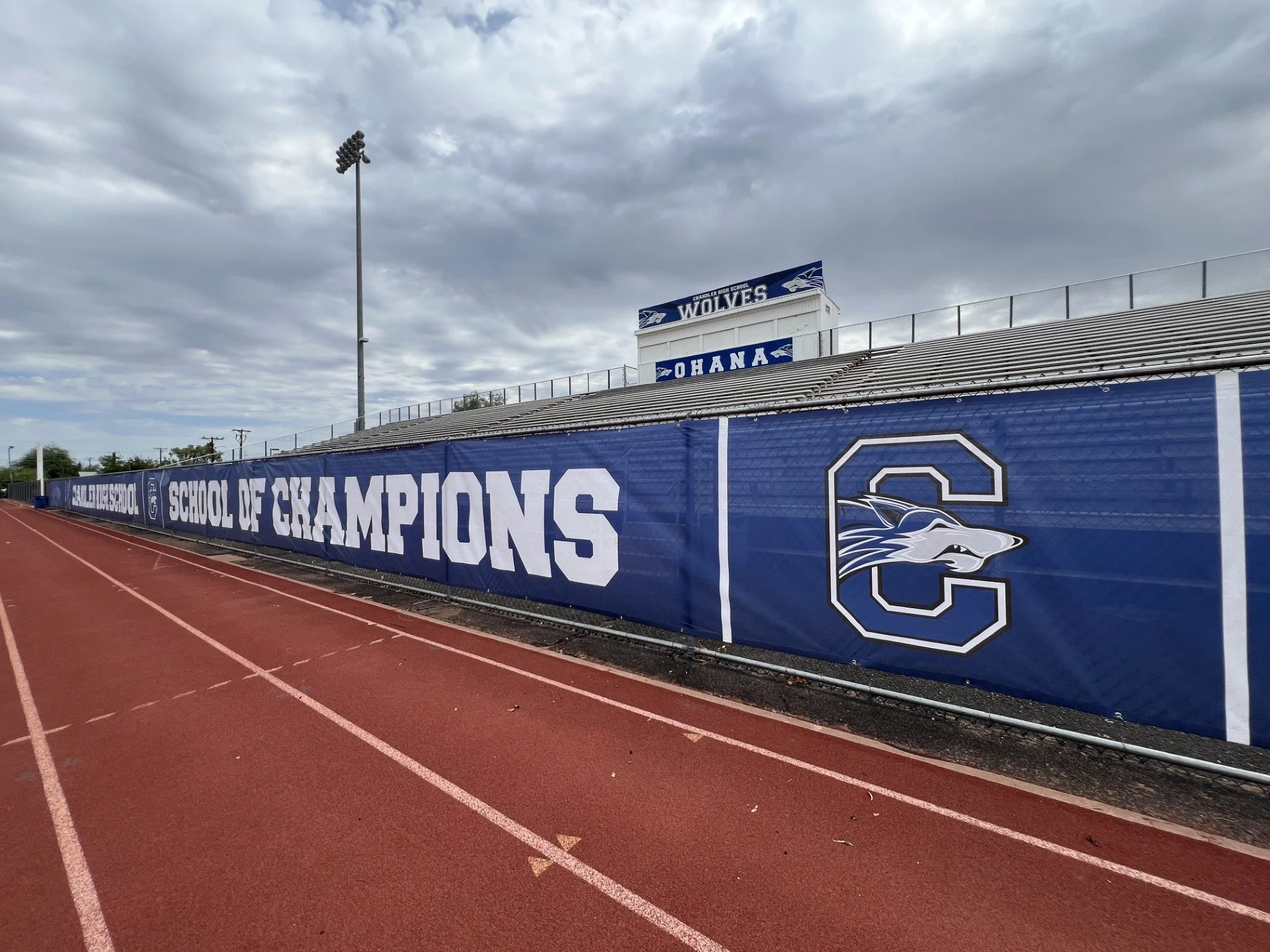 An outdoor sports track with a blue banner that reads 'School of Champions,' and a backdrop of bleachers and a sign that reads 'Chandler High School Wolves.' The sky is cloudy and there is a tall floodlight.