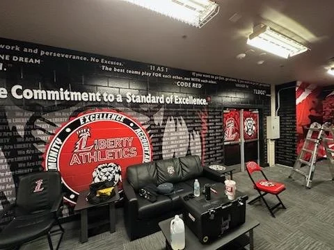 Indoor room with black and red color theme, featuring a large Liberty Athletics logo on the wall, chairs, a small table, water bottles, a ladder, and motivational banners.