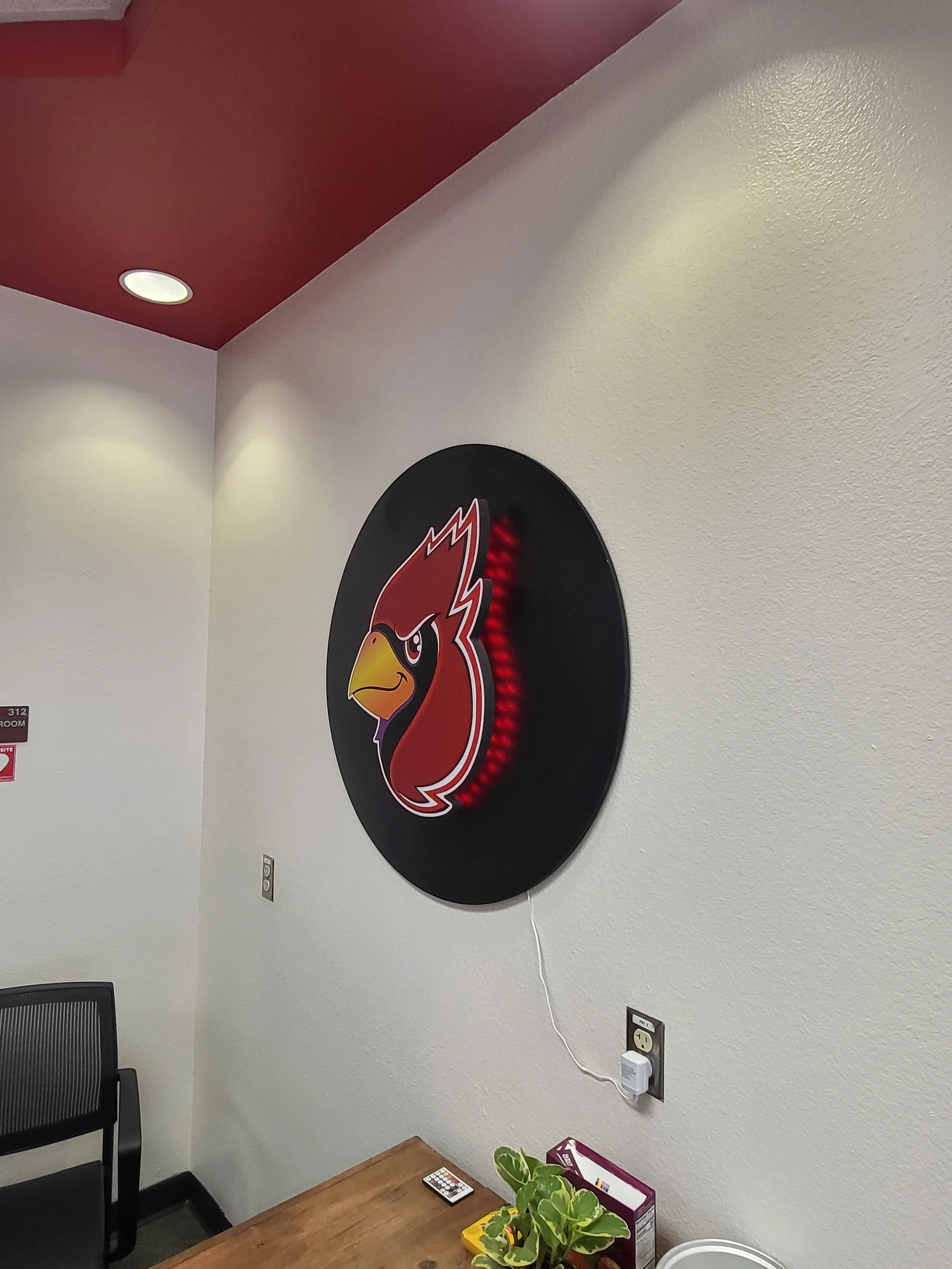 Interior office corner with a white wall, a red ceiling with a recessed light, a black chair, a small table with a remote control and a plant in a purple box, and a wall outlet with a white cable connected.
