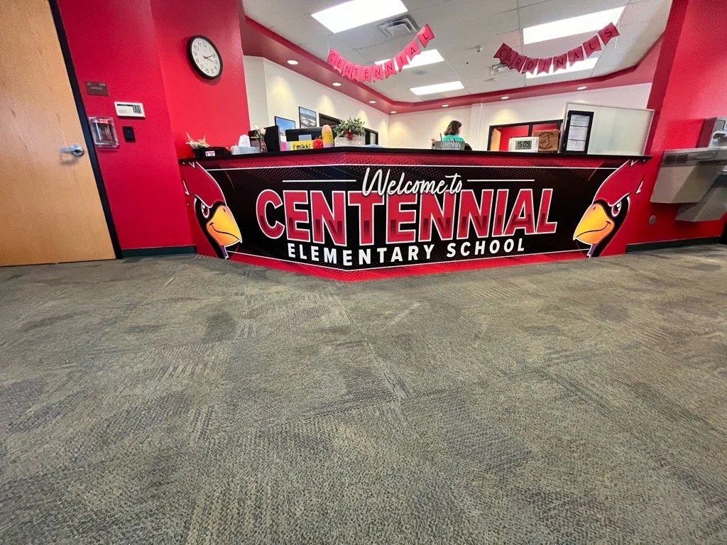Indoor school lobby with a large red and black welcome sign for Centennial Elementary School featuring a cardinal mascot and pendant banner reading 'Celebrating Cardinals'.