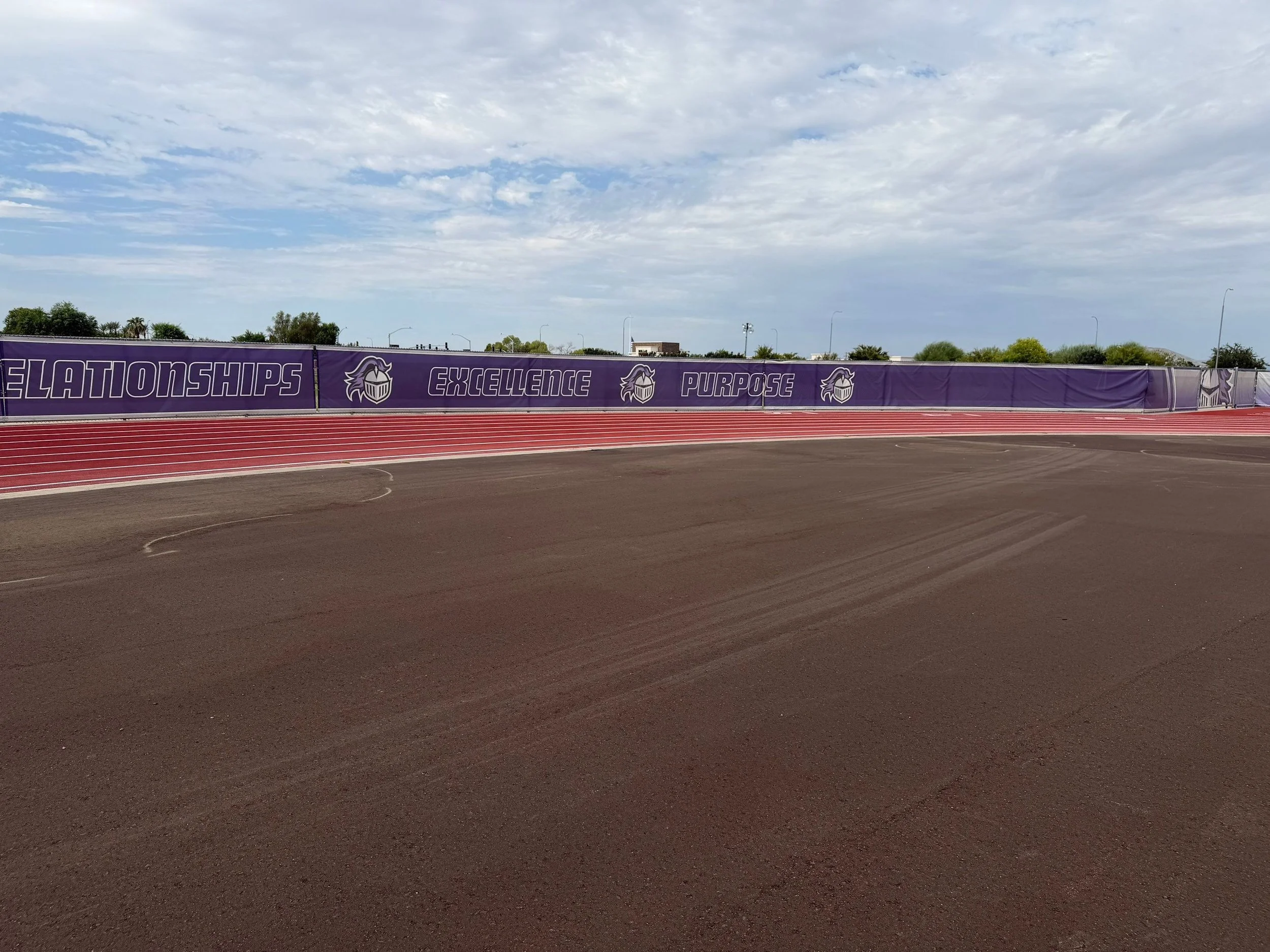 An outdoor running track with a red surface and white lane lines, with a purple banner displaying the words 'Relationships,' 'Excellence,' and 'Purpose,' with a logo, under a partly cloudy sky.