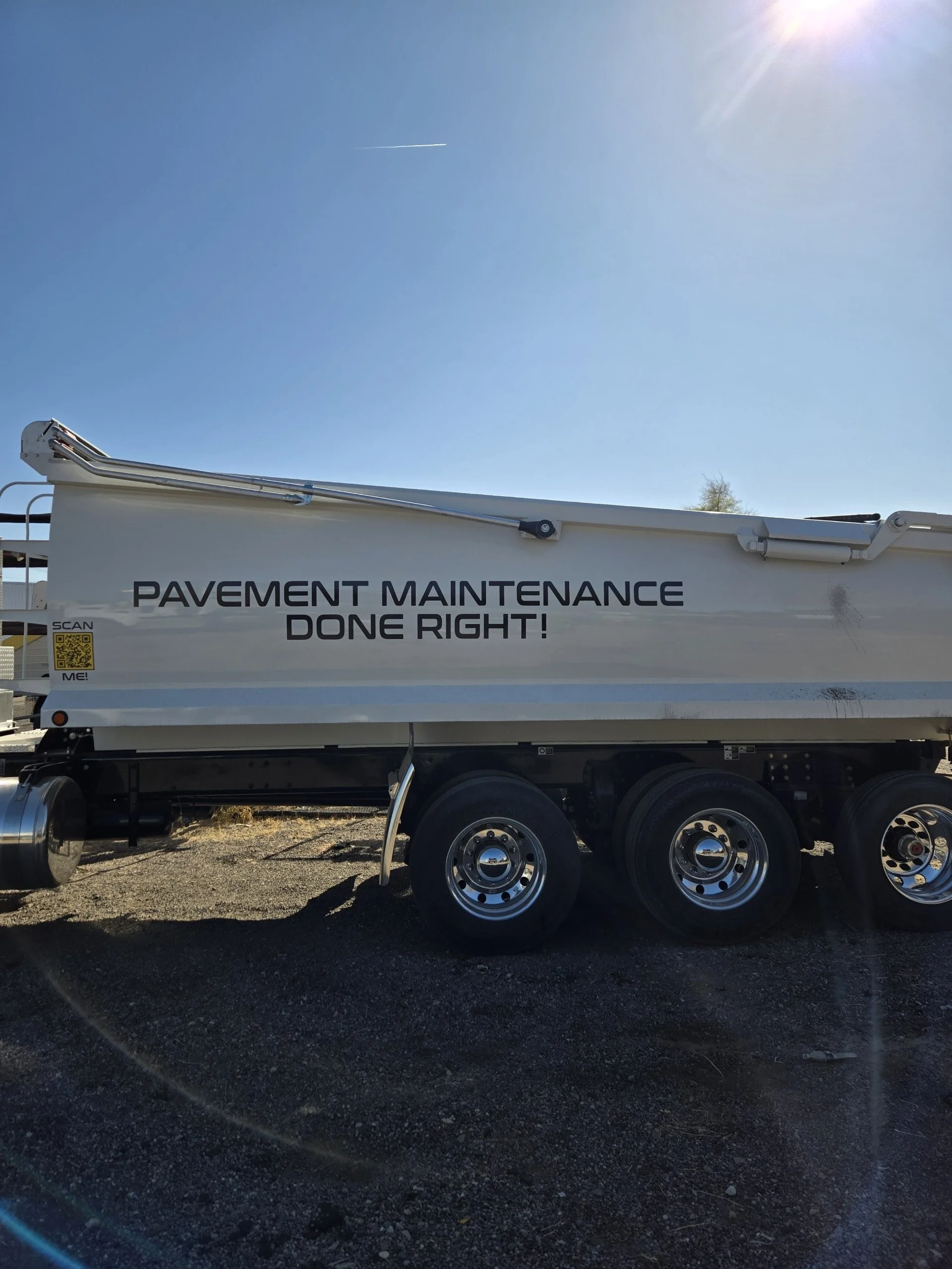 A vehicle with a sign that reads 'Pavement Maintenance Done Right!' parked on a gravel surface under a clear blue sky with the sun shining.