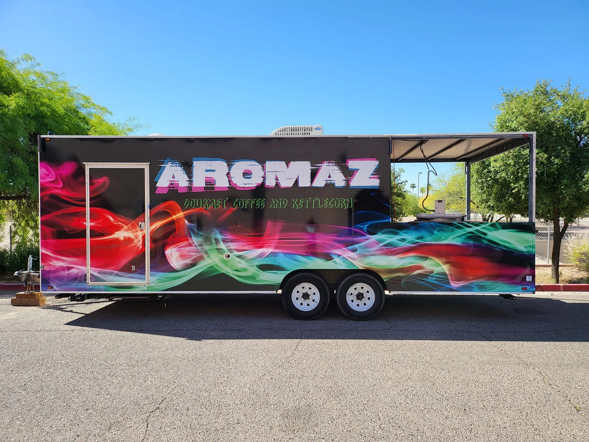 A colorful food truck with vibrant swirling patterns and the text 'AROMAZ' and 'GOURMET COFFEE AND KETTLECORN' on it, parked among trees under a clear blue sky.