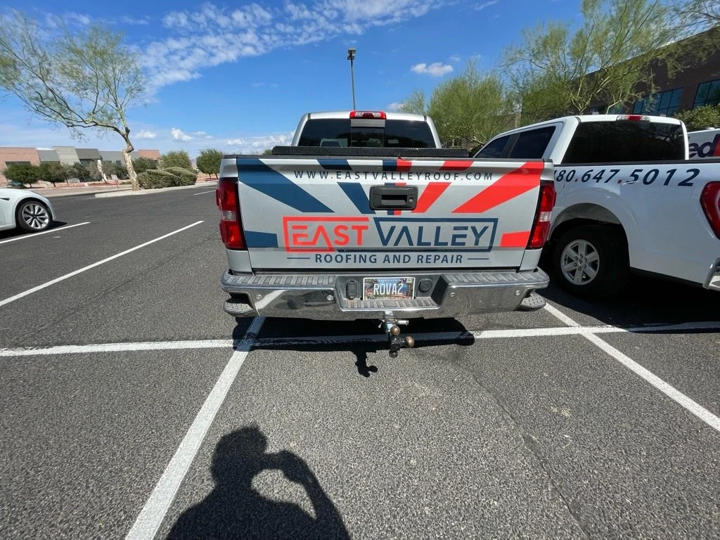 Back of a pickup truck with a logo for East Valley Roofing and Repair, parked in a parking lot with other vehicles and a clear sky.