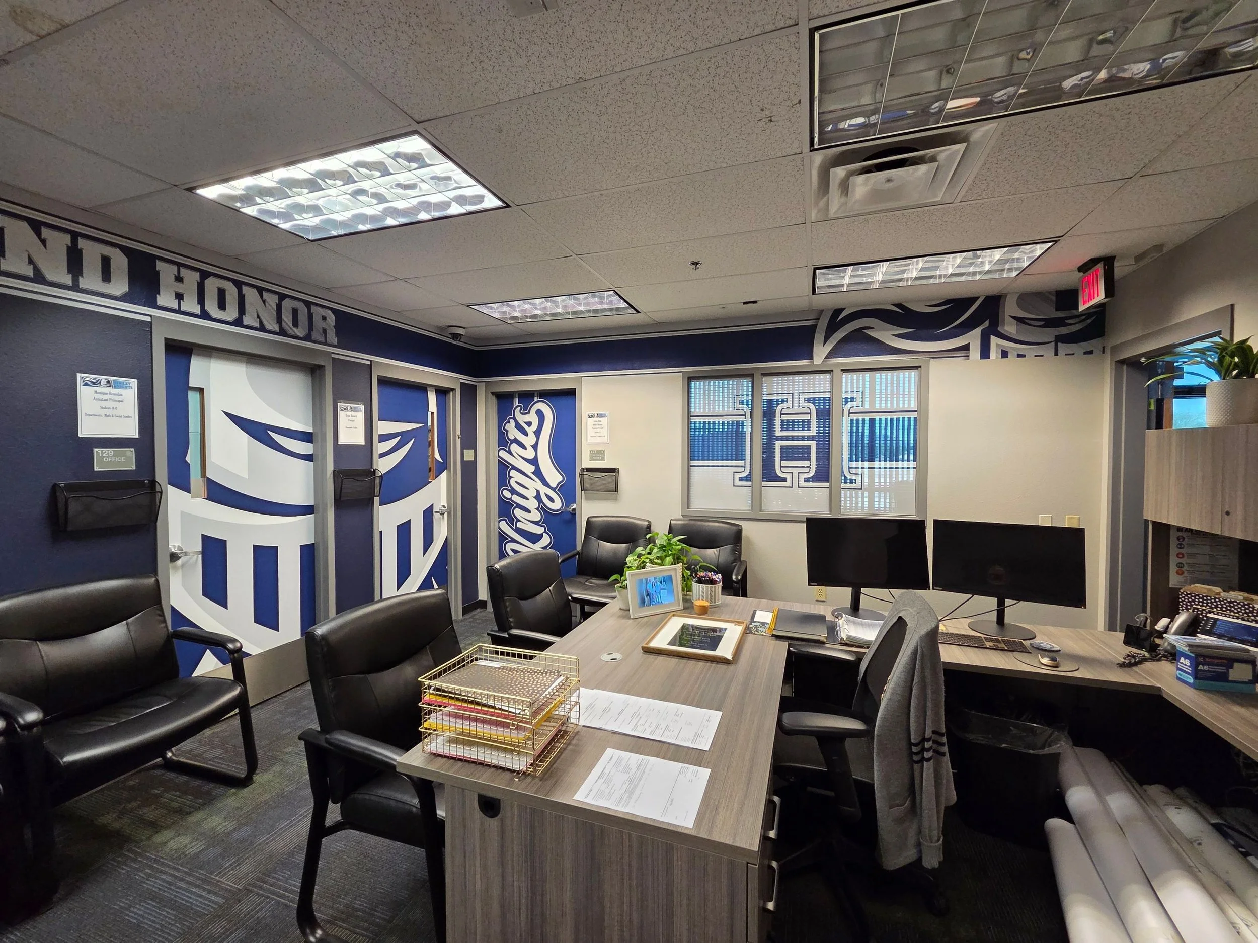 Office waiting room with chairs, a table with papers and office supplies, a large desk, and decorated walls with text and logos.