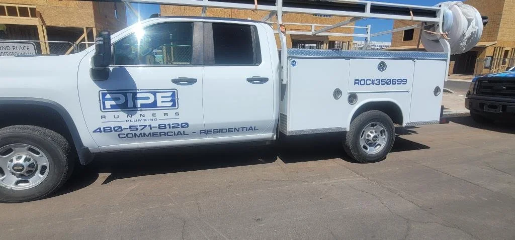 White service truck with 'Pipe Runners Plumbing' logo and contact number, parked on a street with buildings in the background.