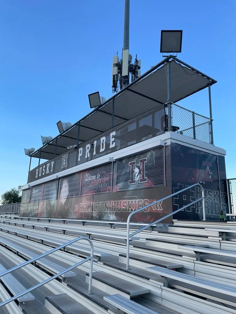 Empty football bleachers with Husky Pride signage, a press box displaying 'HAMILTON MUSKIES', and a large scoreboard or screen at the top, under a clear blue sky.