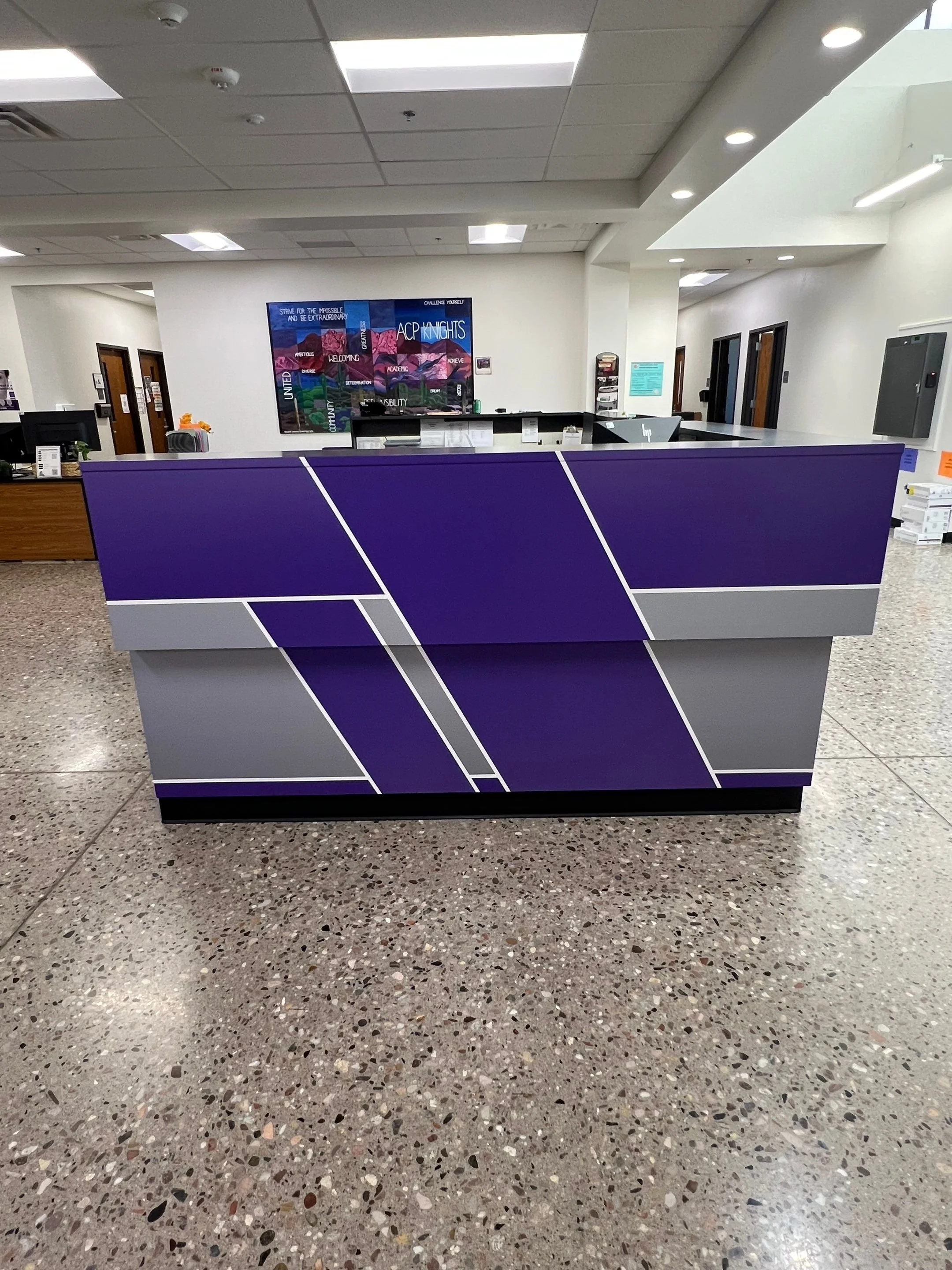 View of a reception desk in an office or educational building with purple and grey geometric design, computer monitors, and a wall-mounted colorful poster with mountains and text.