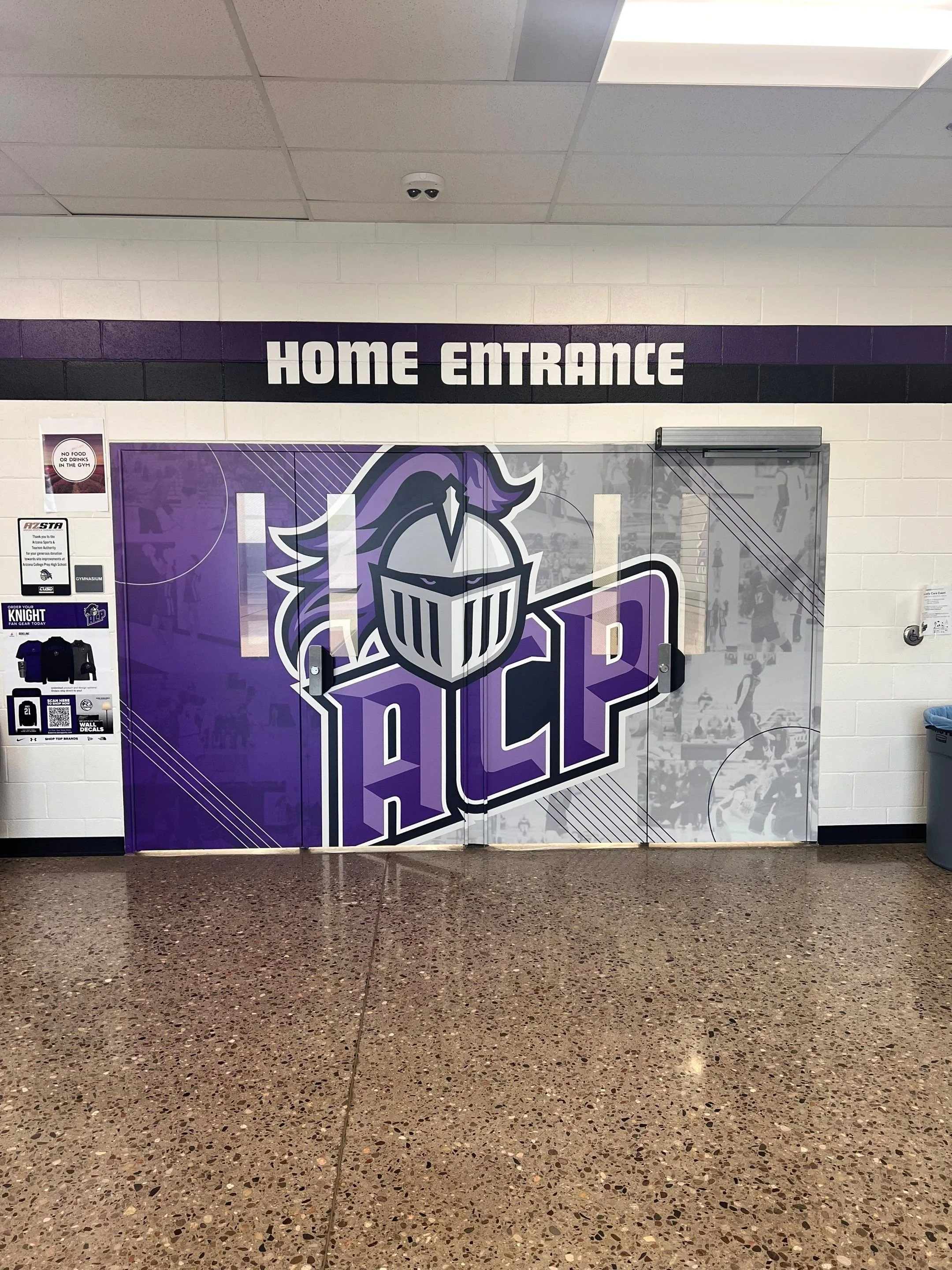Entrance door with a sign above it that reads 'Home Entrance' in white letters on a black background, and a purple and white athletic logo with a knight helmet on the door, located inside a school or gymnasium.