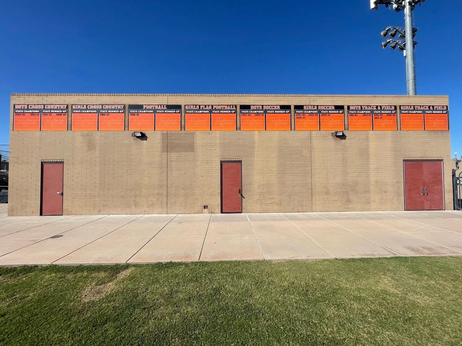 A beige brick building with eight orange and black boards on the upper part displaying different sports achievements, two red doors, two black lights, and a tall metal light pole against a clear blue sky.