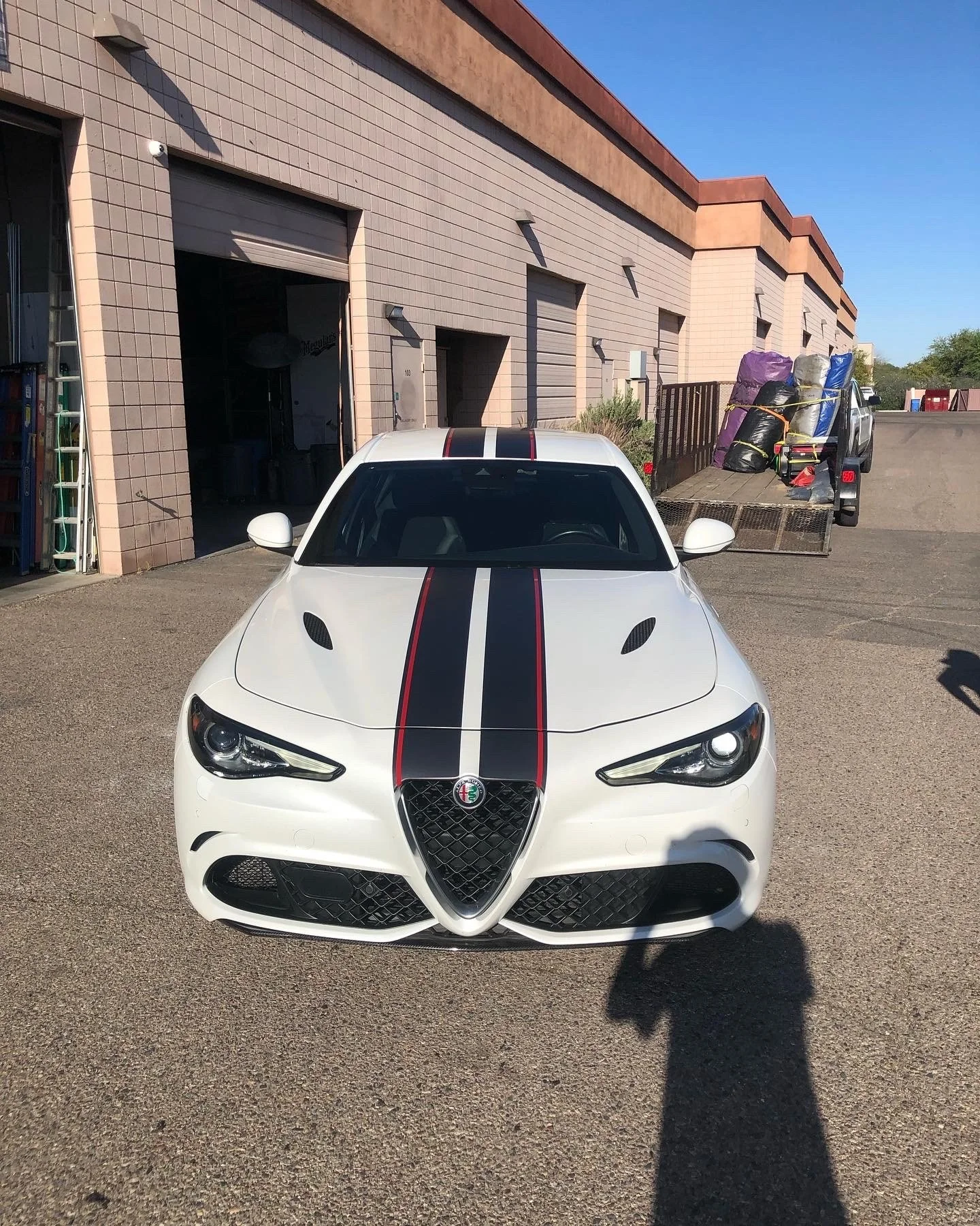 White sports car with two black and red racing stripes on the hood, parked outside a warehouse with a partially open garage door, and a flatbed truck loaded with bags and equipment in the background.
