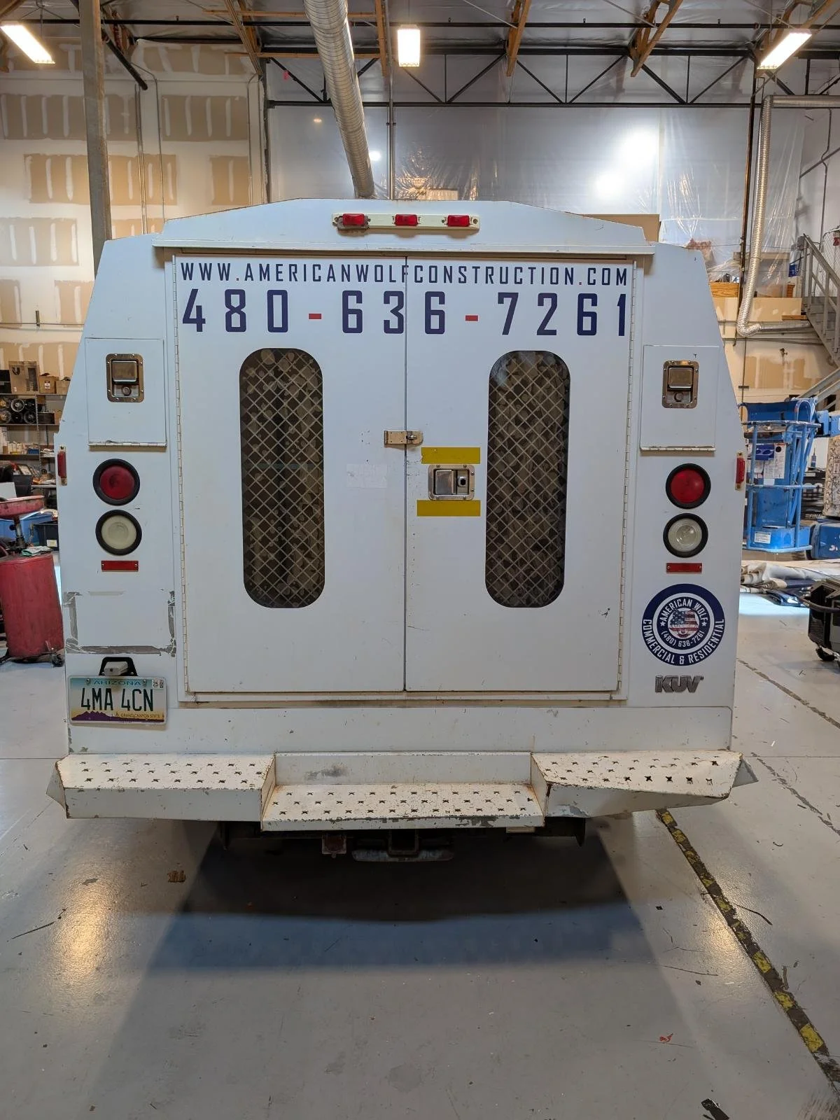 Back of a white utility vehicle with website, phone number, and company logo, parked inside a warehouse with tools and equipment around.