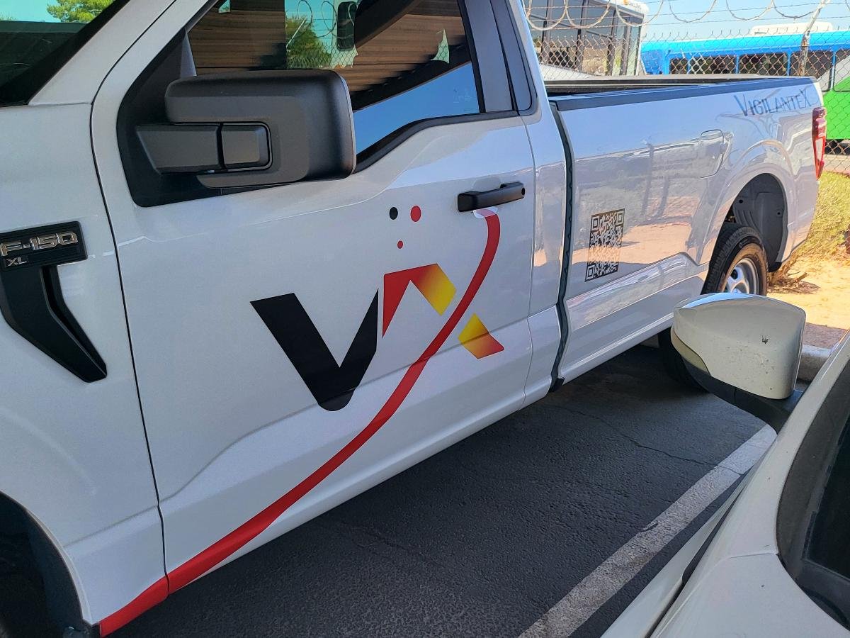 A white pickup truck with the company's logo and design on the side, parked in a parking lot near a chain-link fence and other vehicles.