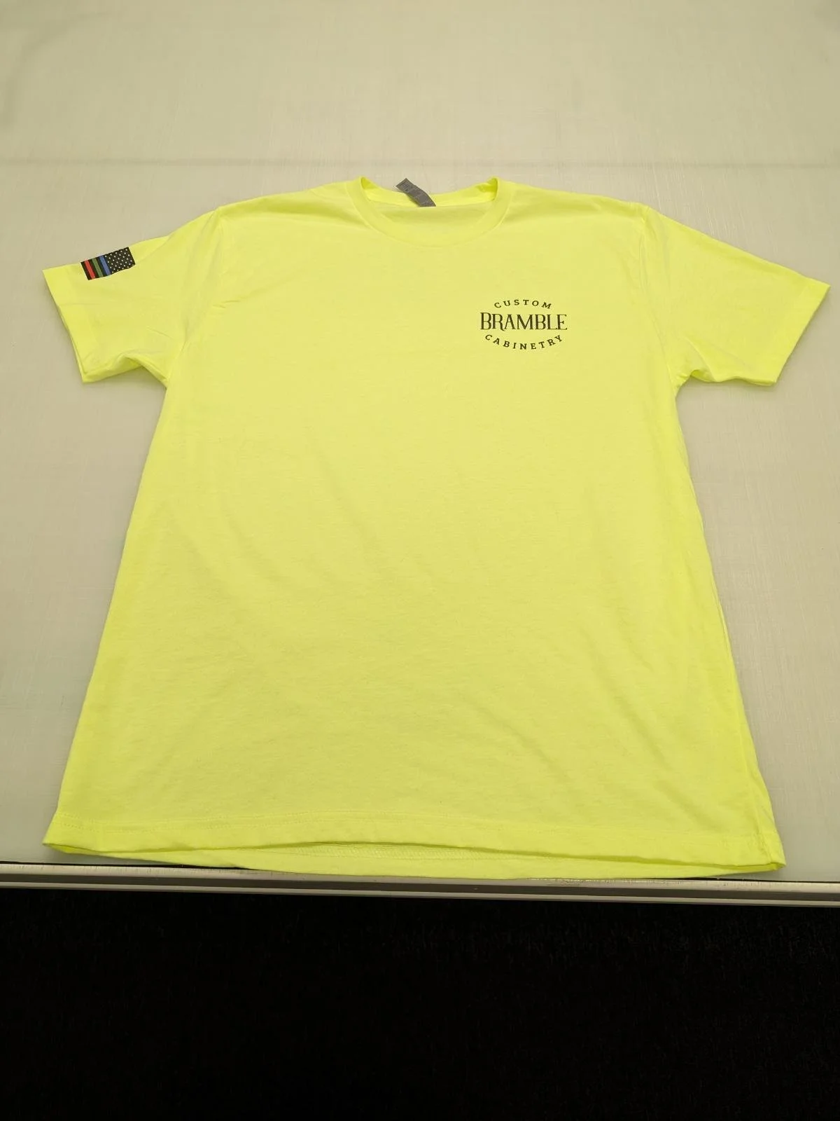 Yellow T-shirt with a flag patch on the sleeve and a logo on the front that reads 'Custom Bramble Cabinetry'.