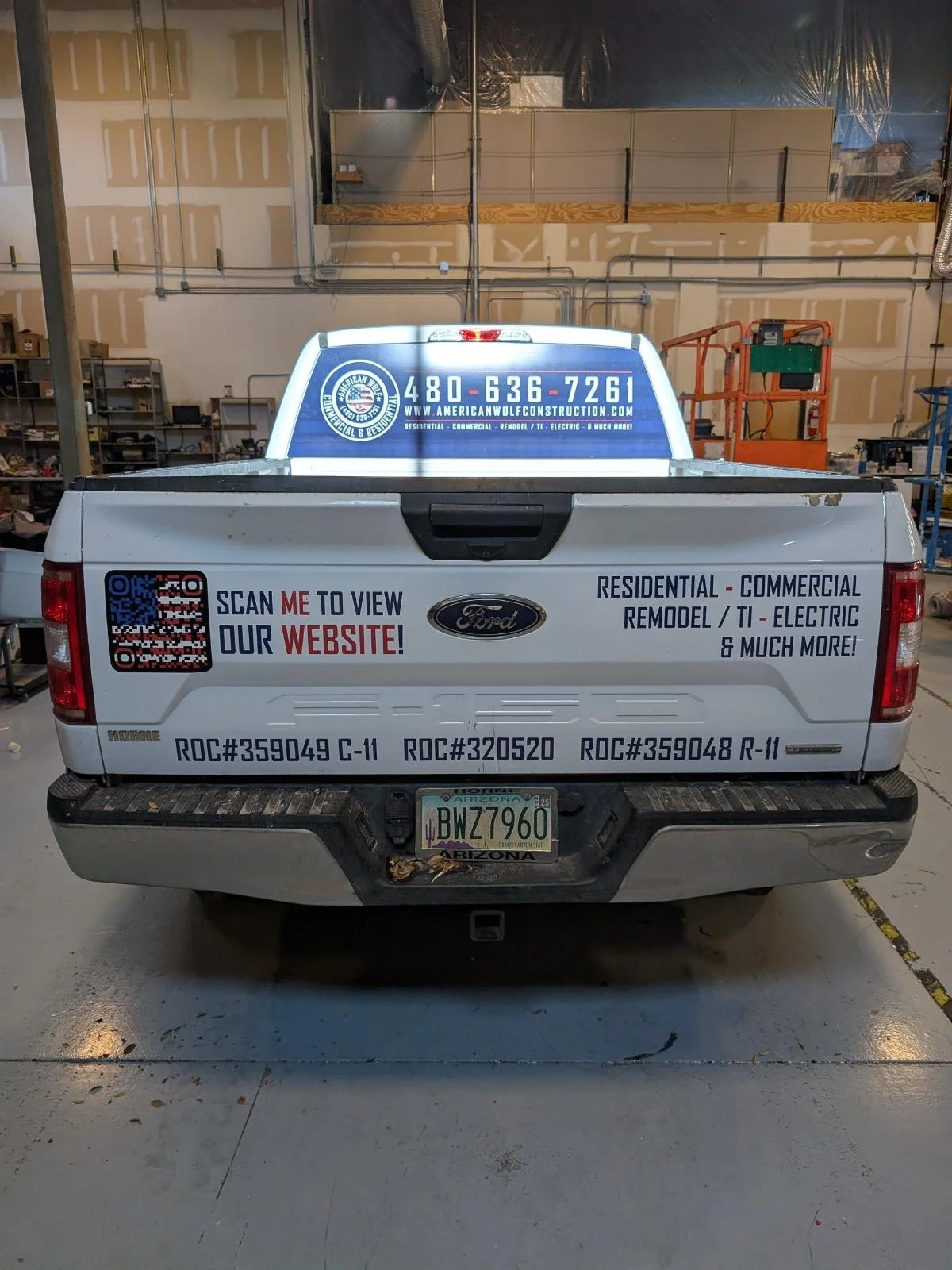 The rear of a white Ford pickup truck inside a workshop or garage with various tools and equipment. The truck has automotive business advertisements on the tailgate, including a QR code, a website link, and contact information. It also displays sever