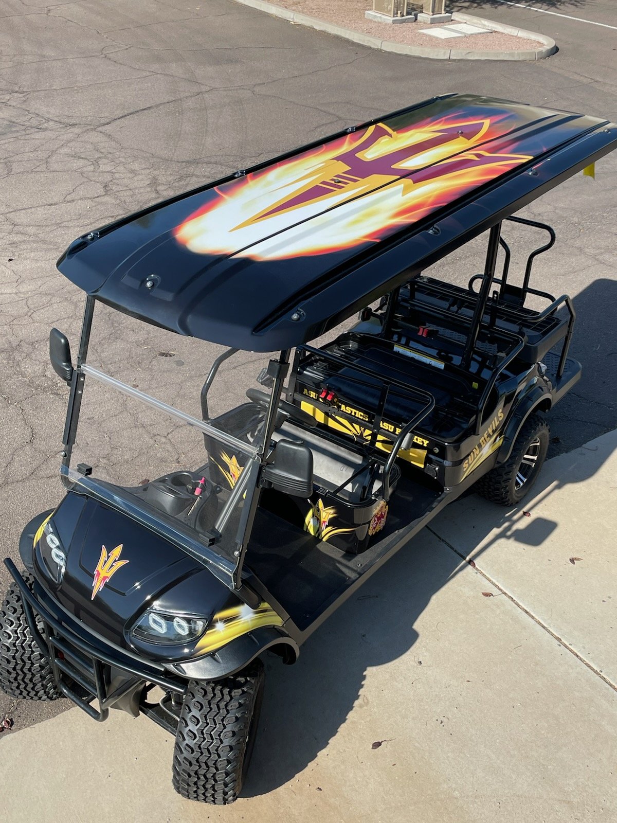 A custom golf cart with a black body and yellow flaming graphics, featuring a large graphic on the roof of a yellow arrow with purple outlines. The cart has a black windshield, large off-road tires, and is parked on a paved surface near a sidewalk.