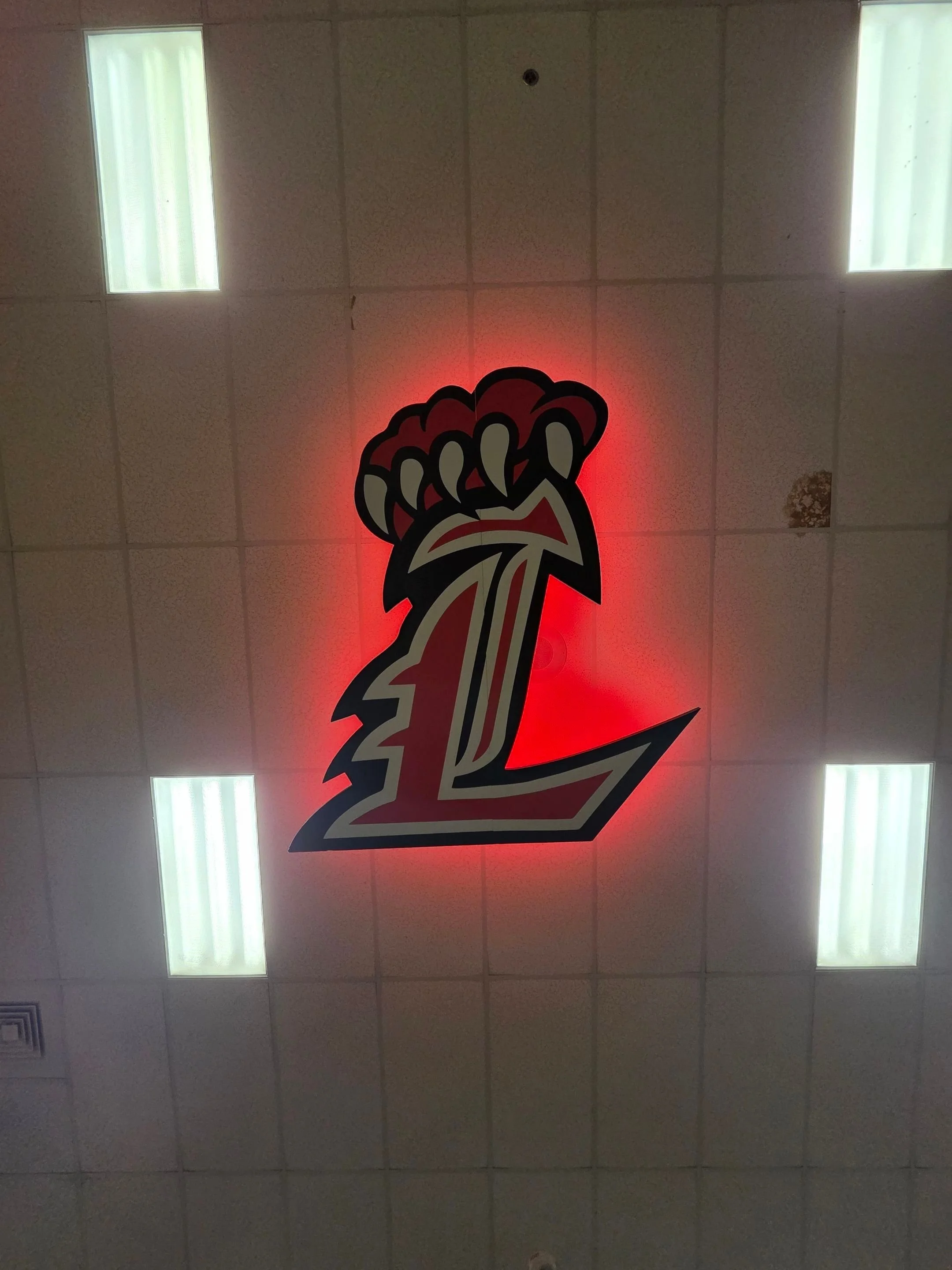 Ceiling with a large illuminated logo of the Kansas City Chiefs football team, featuring a red arrowhead and a stylized arrow, with fluorescent lights and ceiling tiles visible.