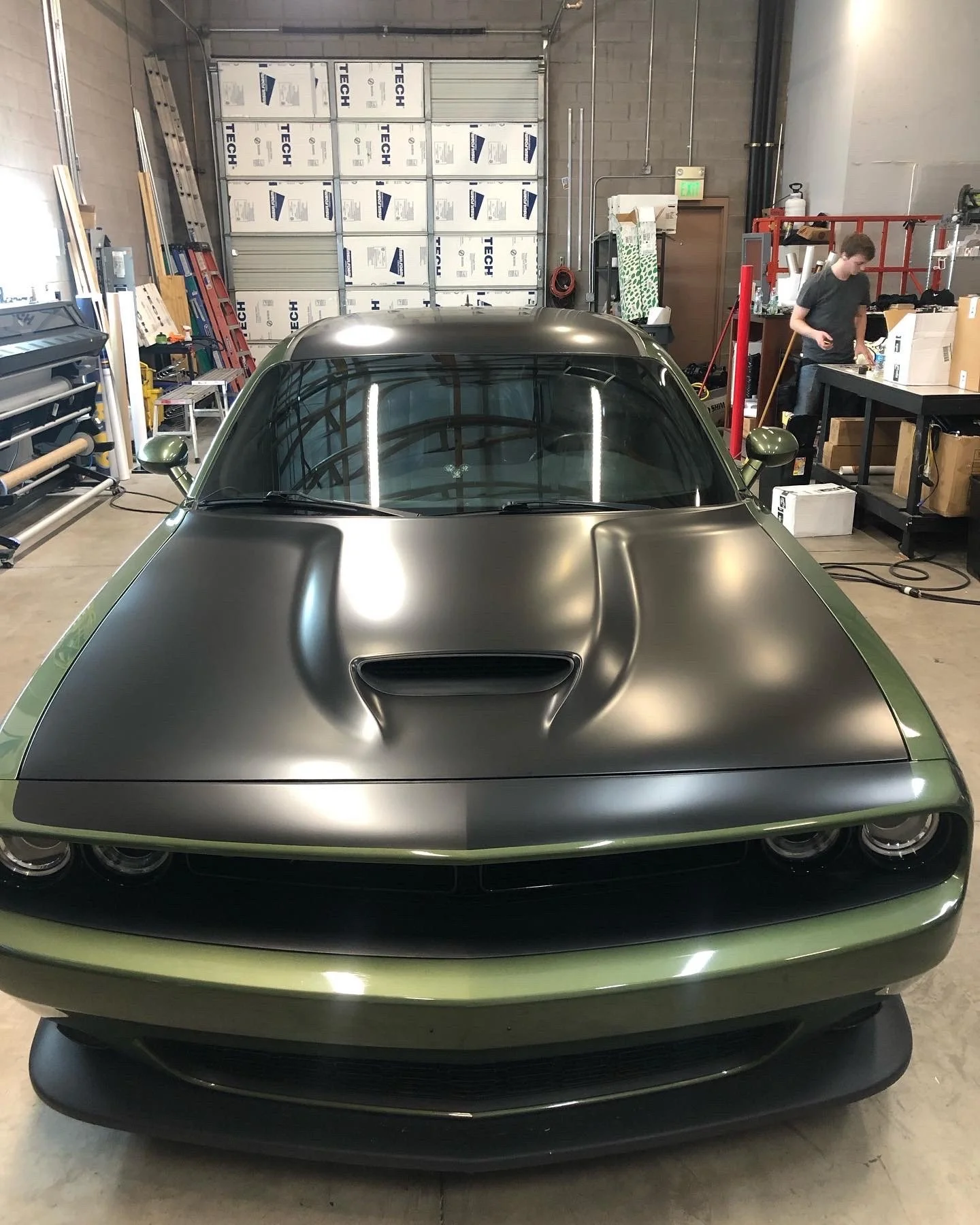 Front view of a sleek, matte black and olive green vintage sports car inside a workshop.