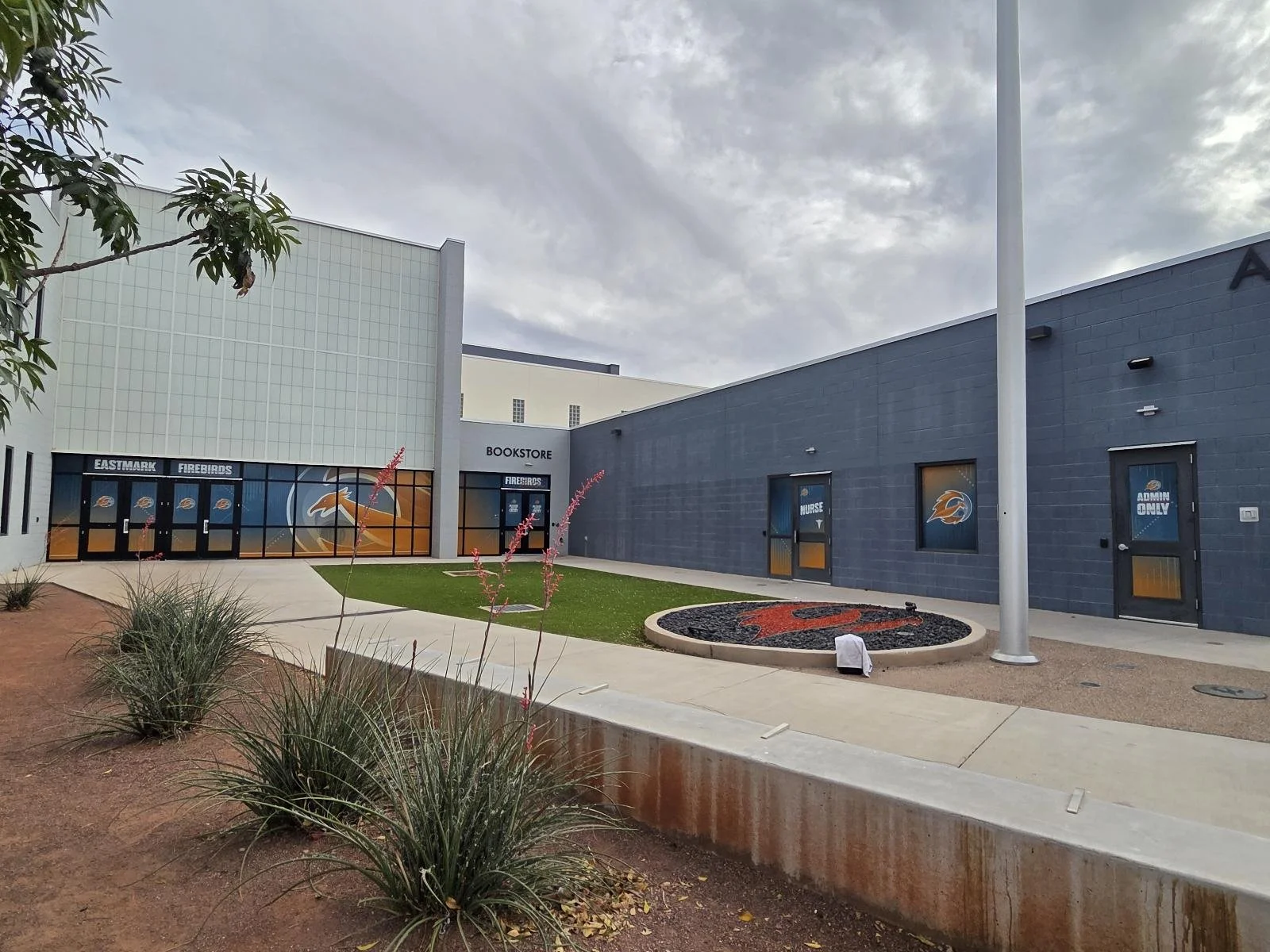 Exterior view of a sports arena with a blue and white facade, featuring windows with team logos, a small grass area, and a sign reading 'BOOKSTORE'; cloudy sky overhead.