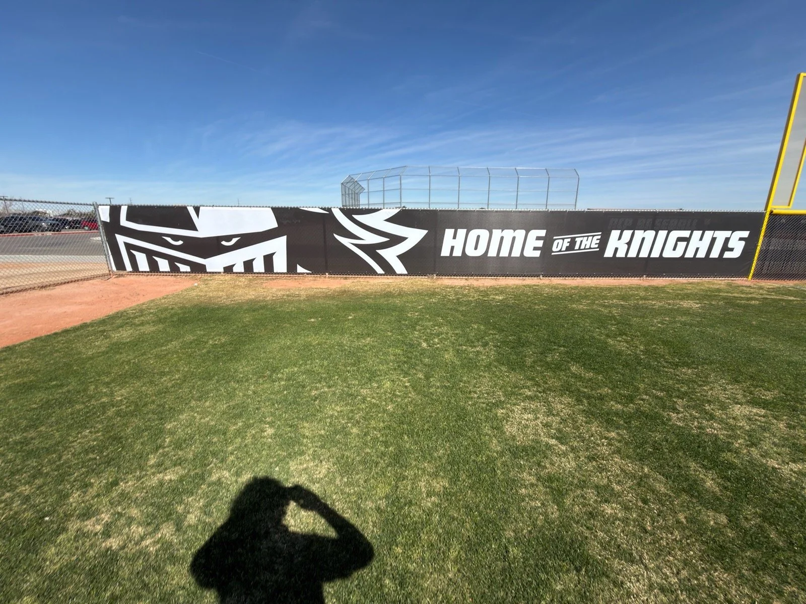 A baseball field with a fence and a black banner displaying a knight's helmet logo and the words 'Home of the Knights' in white letters.
