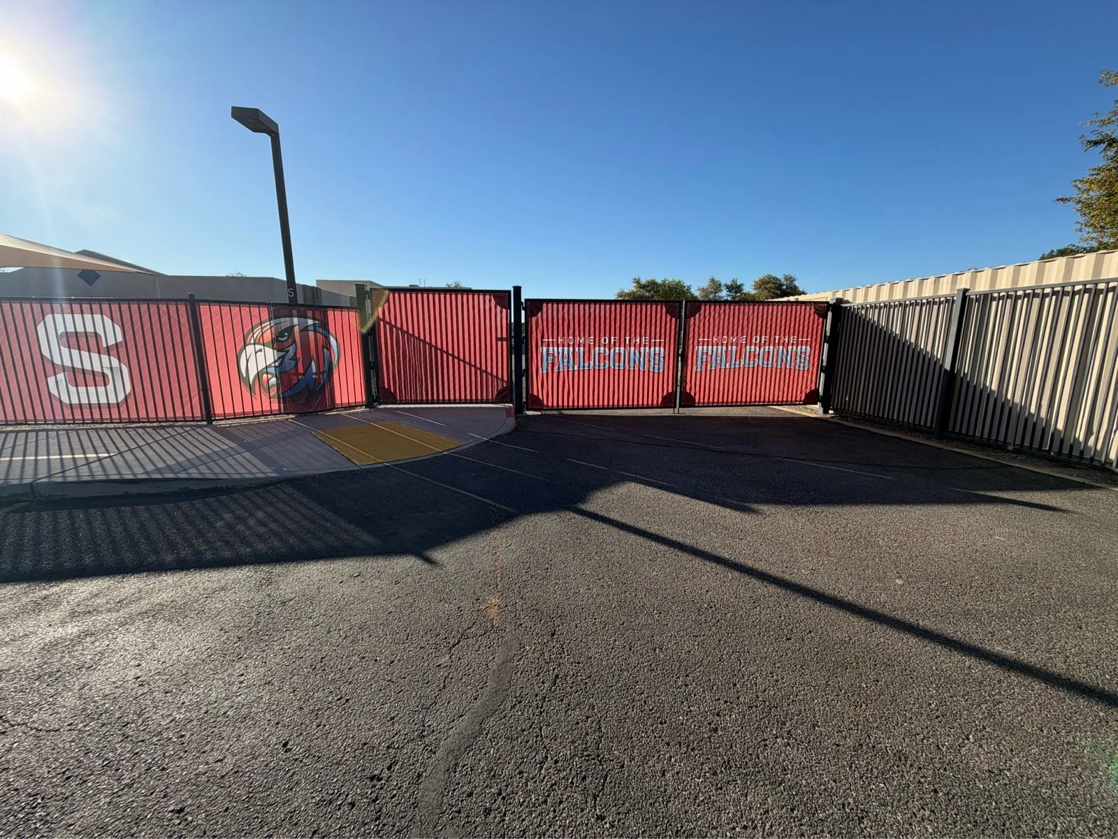 A black metal gate with red banners featuring a falcon mascot and the words "Home of the Falcons" on a sunny day, with a clear blue sky and streetlight in the background.