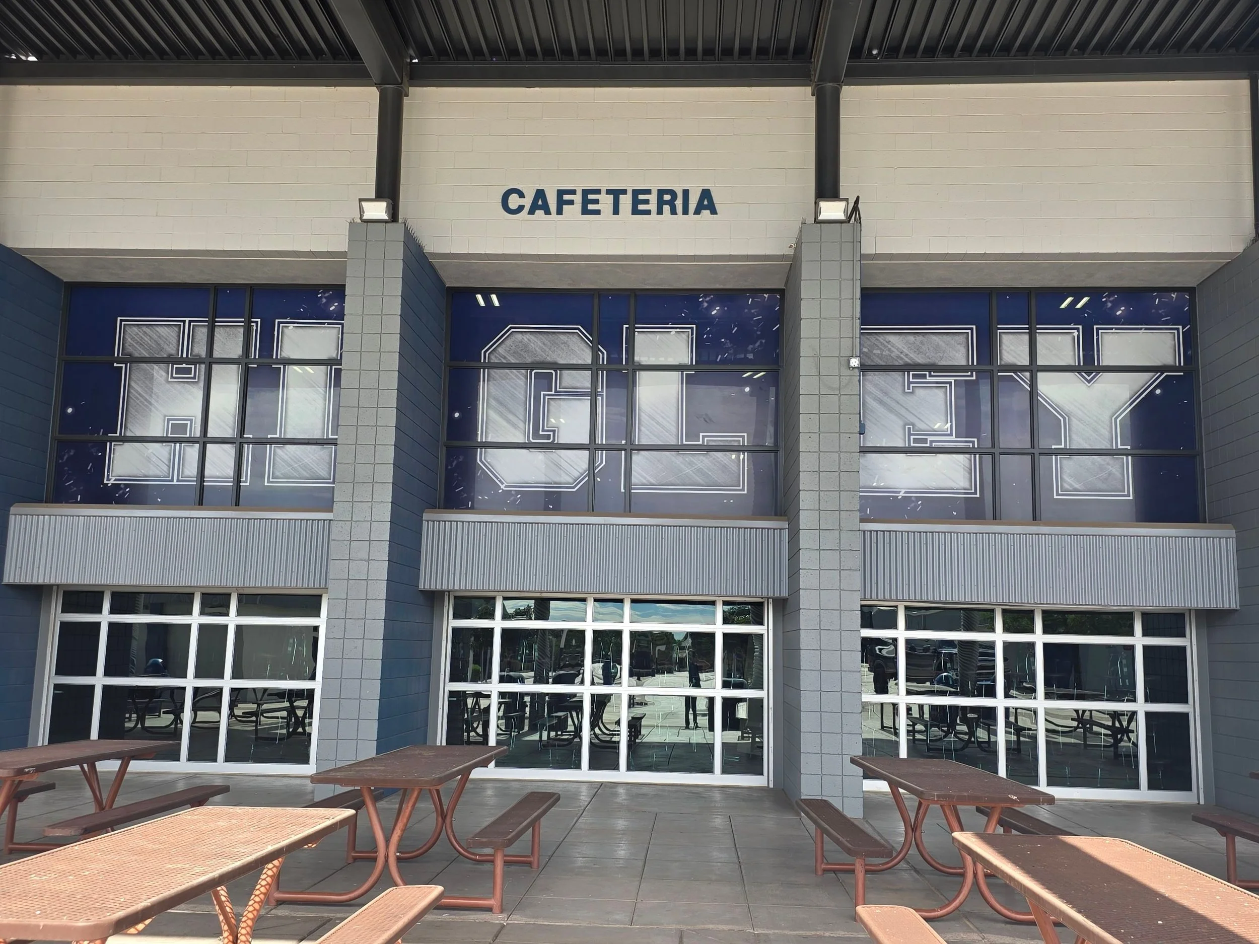 Exterior view of a cafeteria with large windows, a sign that says "CAFETERIA" above the entrance, and outdoor seating with wooden benches.
