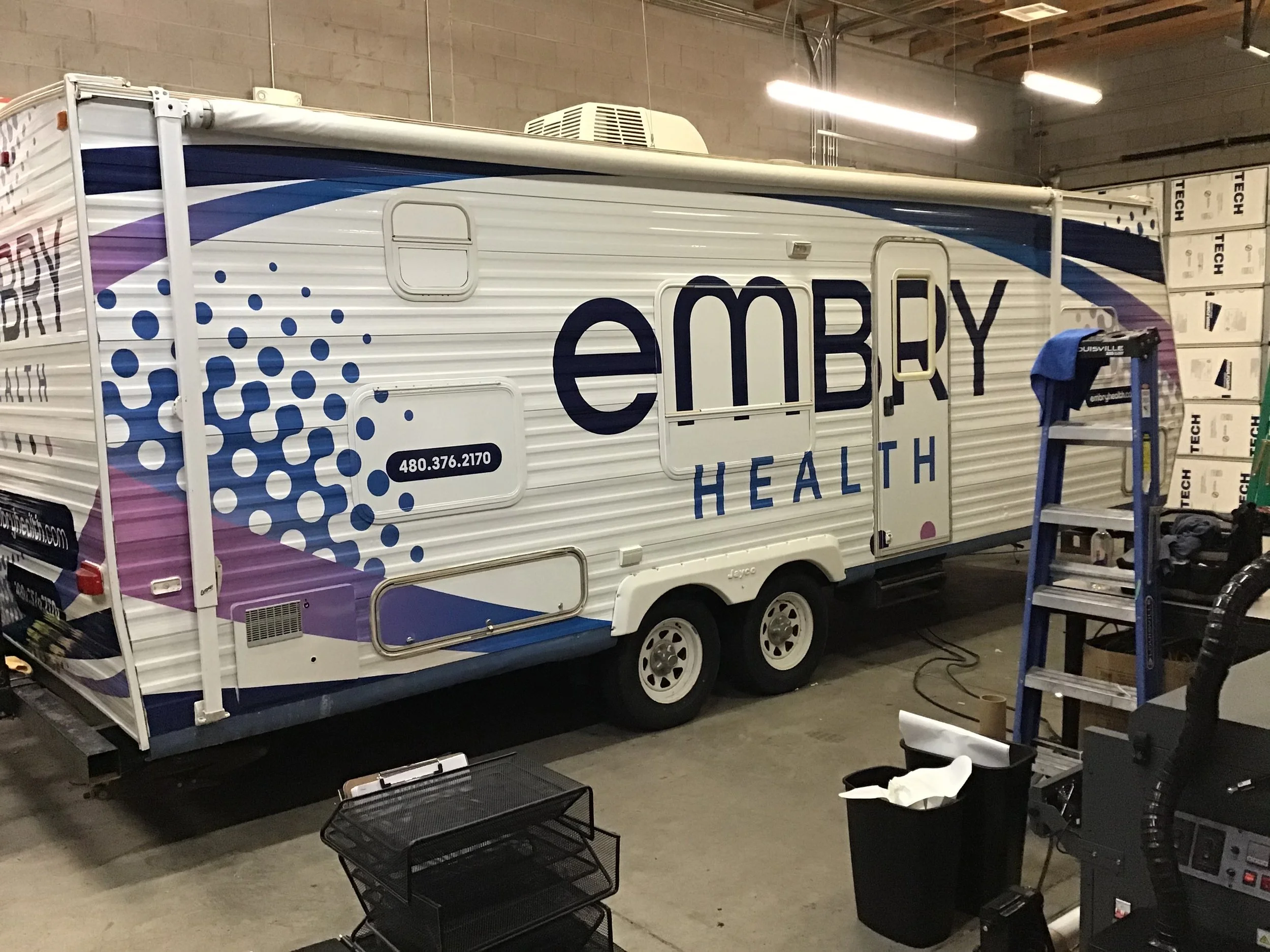 A mobile health clinic trailer inside a warehouse with boxes and equipment around, branded with 'Emergency Health' and contact number.